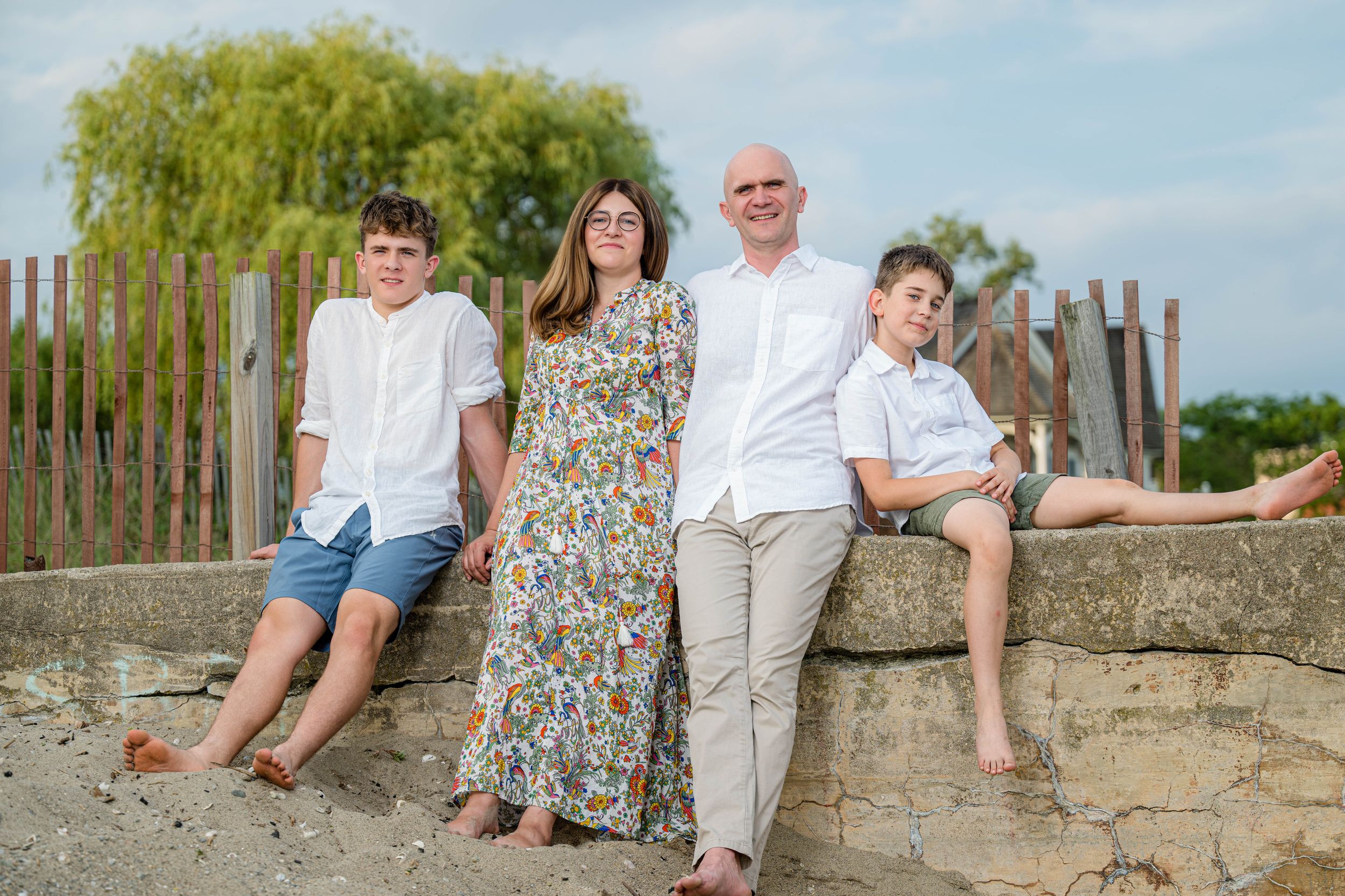 Family of four posing outdoors on a stone wall, wearing white and floral outfits, with greenery in the background.