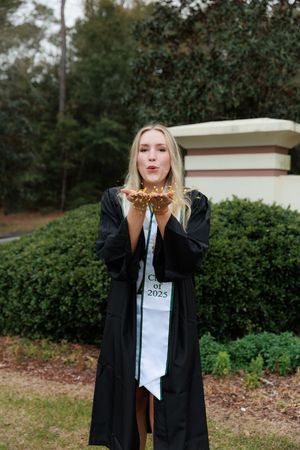 Blonde graduate in black cap and gown blows glitter from hands, wearing Class of 2025 stole.