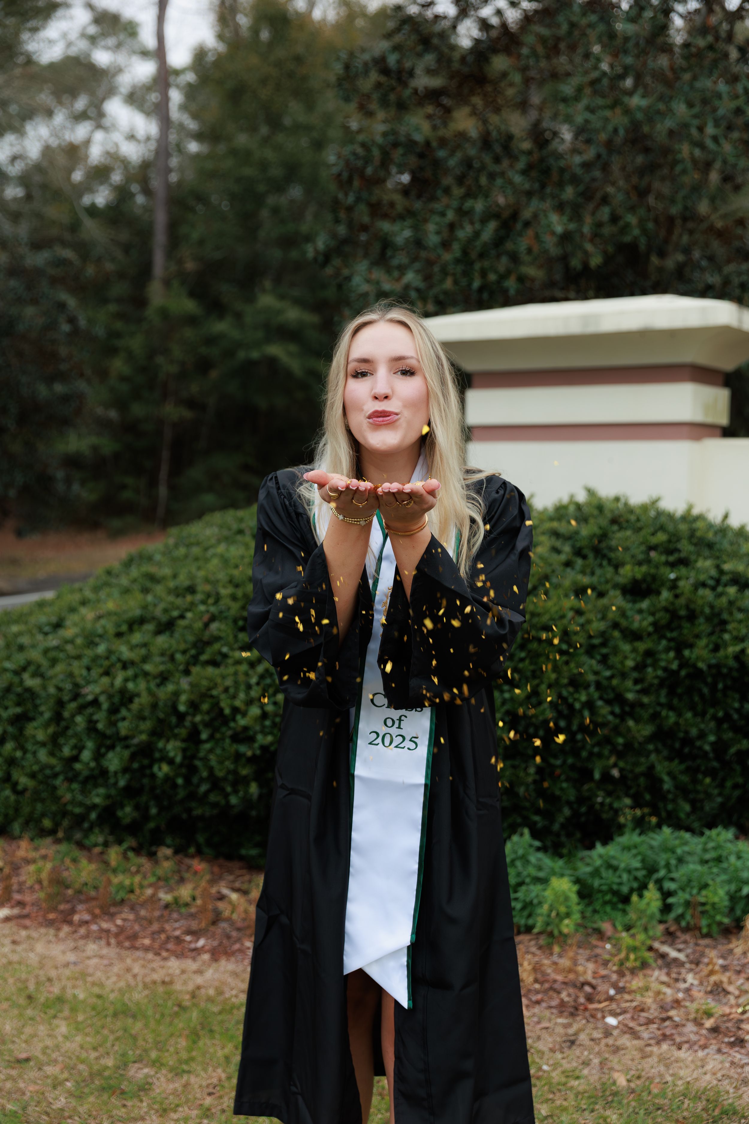 Blonde woman in black graduation gown blowing gold confetti, wearing Class of 2025 sash, outdoors.