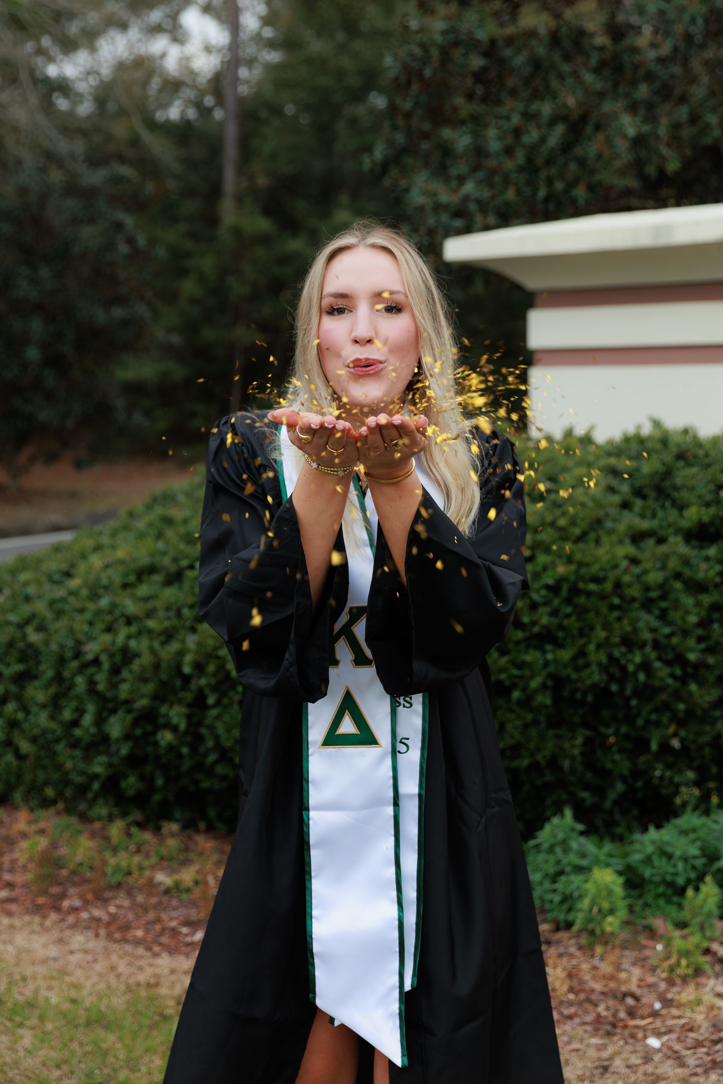 Blonde woman in graduation gown and sorority sash blowing gold confetti outdoors.