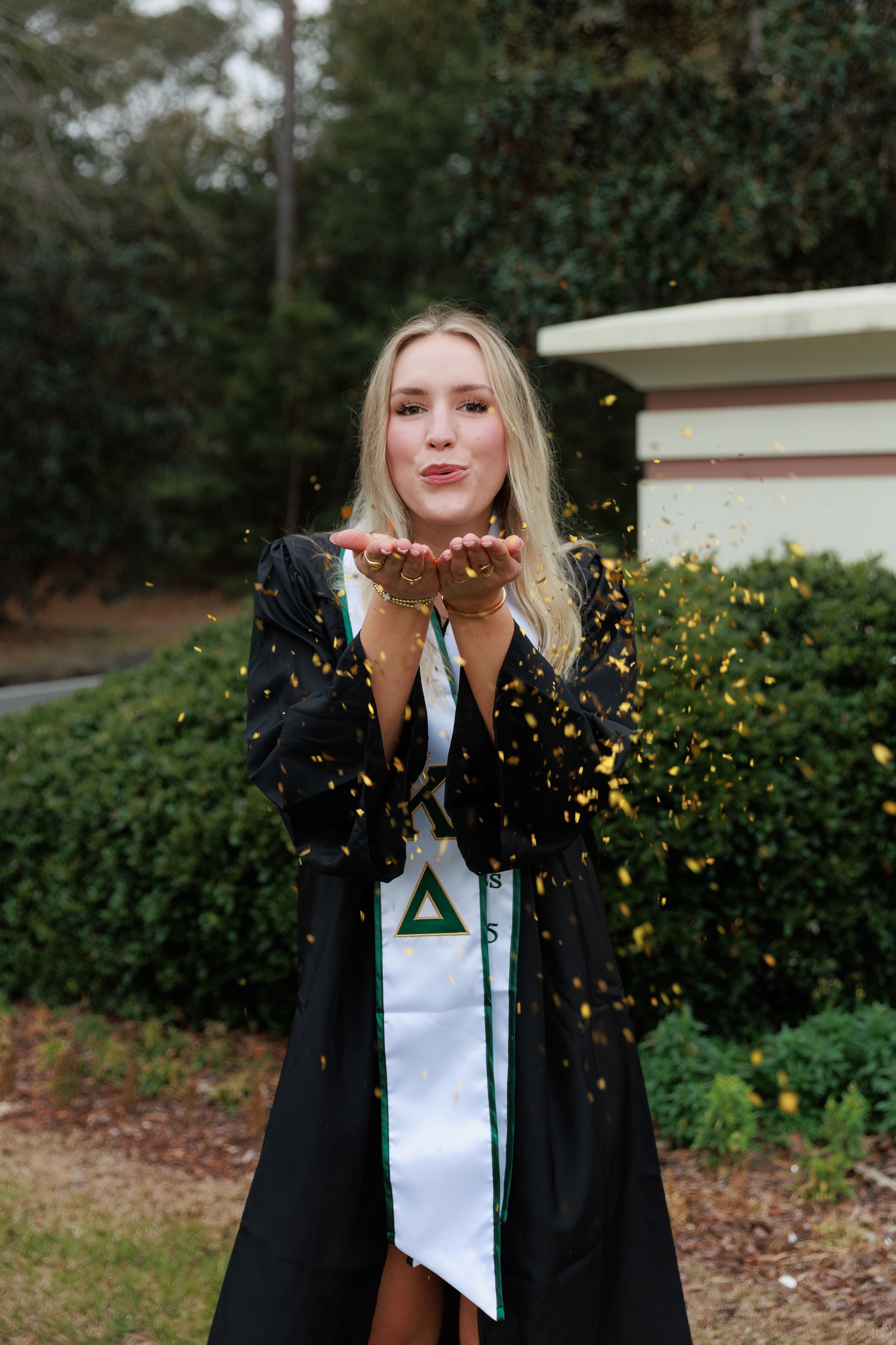 Blonde graduate in black gown blowing gold confetti with Greek letter stole outdoors near greenery.