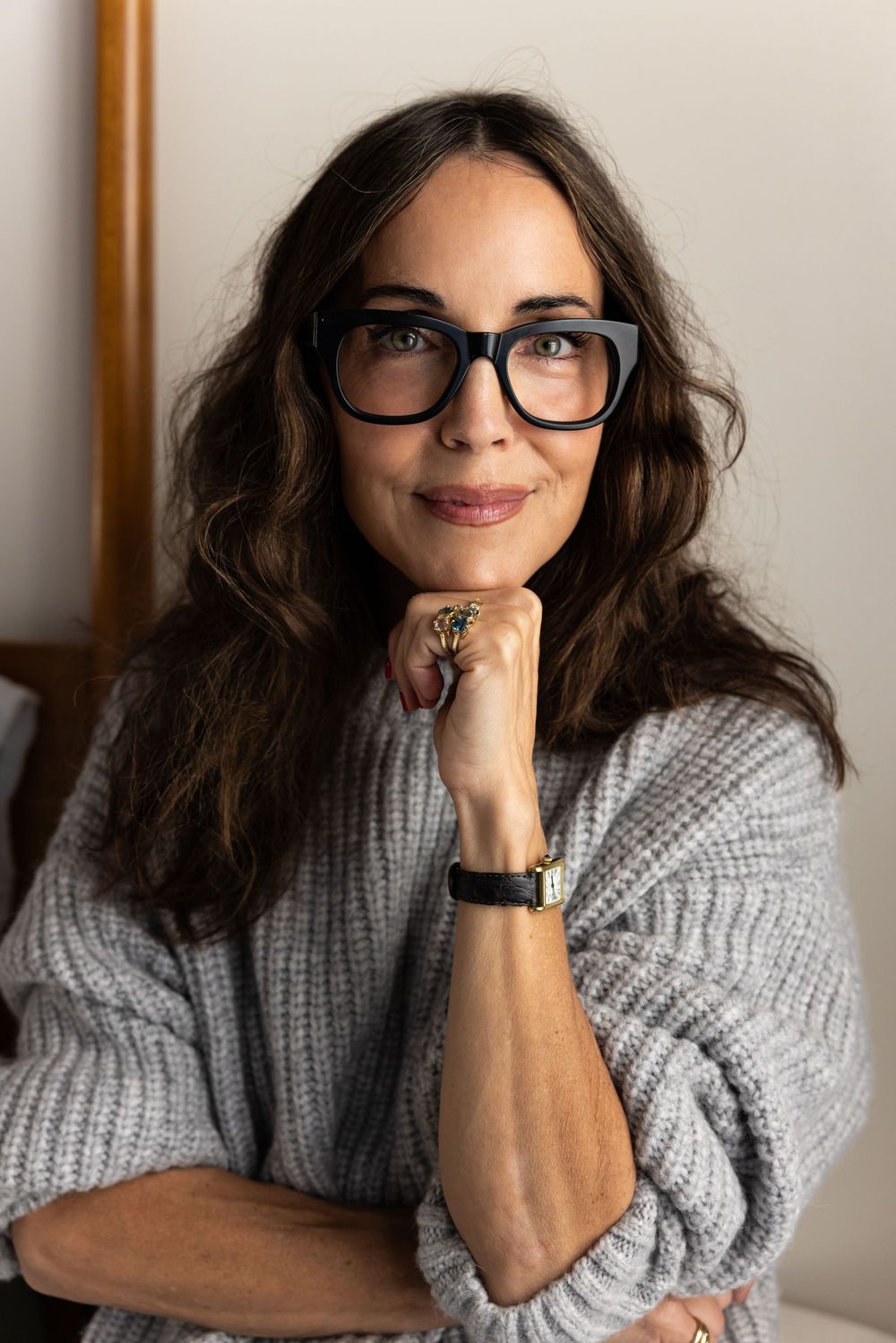 Brunette woman in gray knit sweater and black glasses poses thoughtfully indoors.