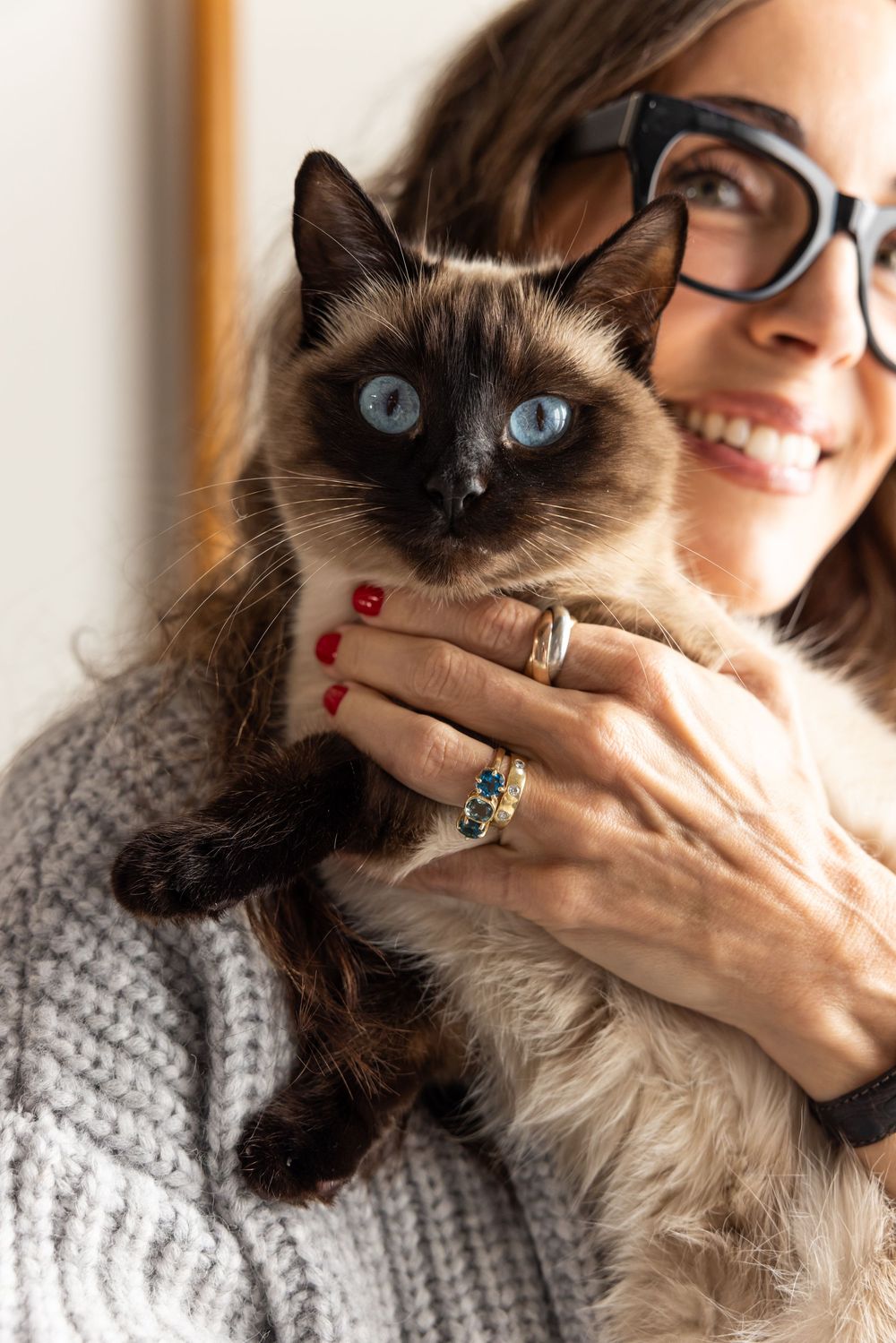 Smiling woman in gray sweater holds a Siamese cat with striking blue eyes.