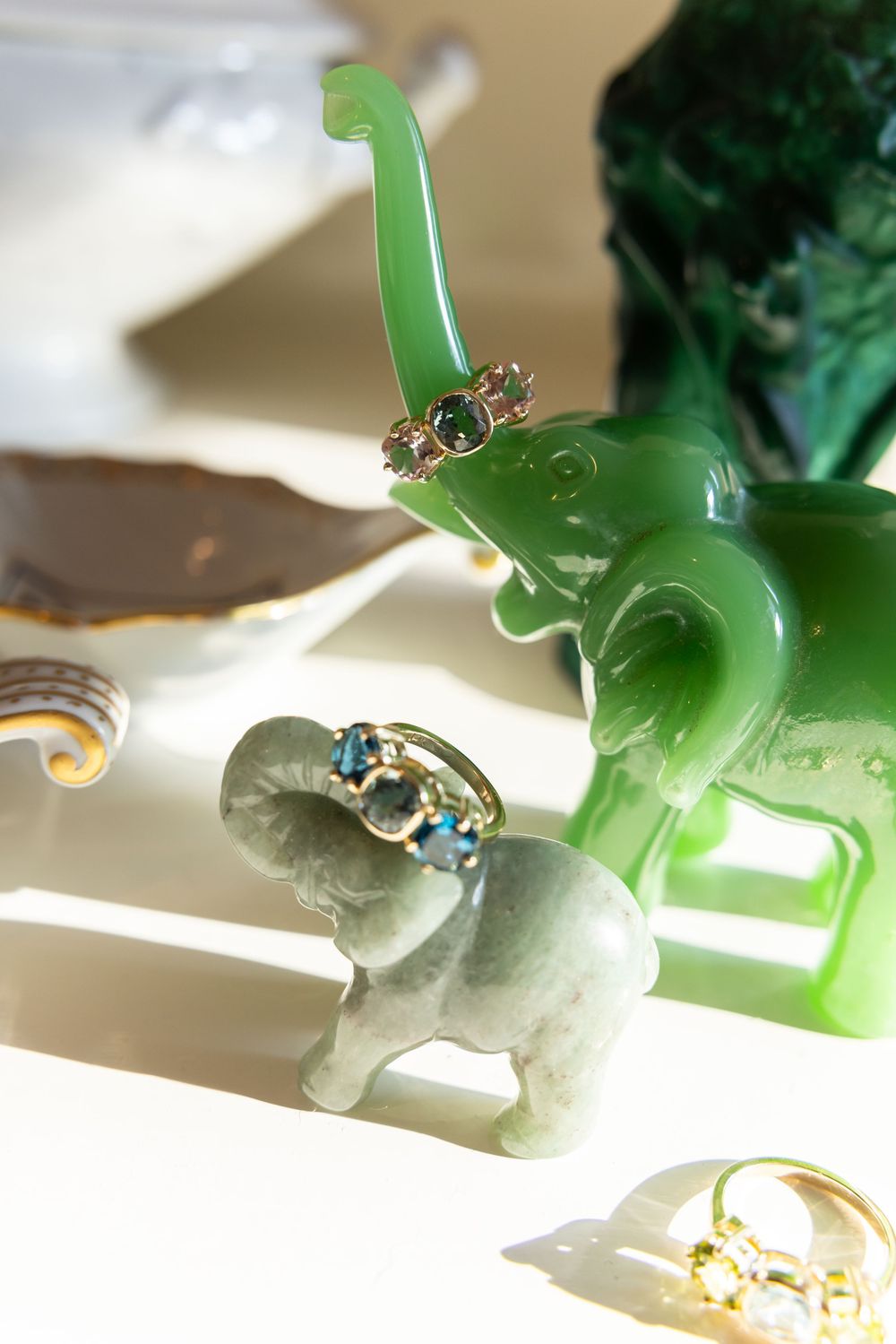 Two jade elephant figurines displaying gold and gemstone rings on a bright white surface.