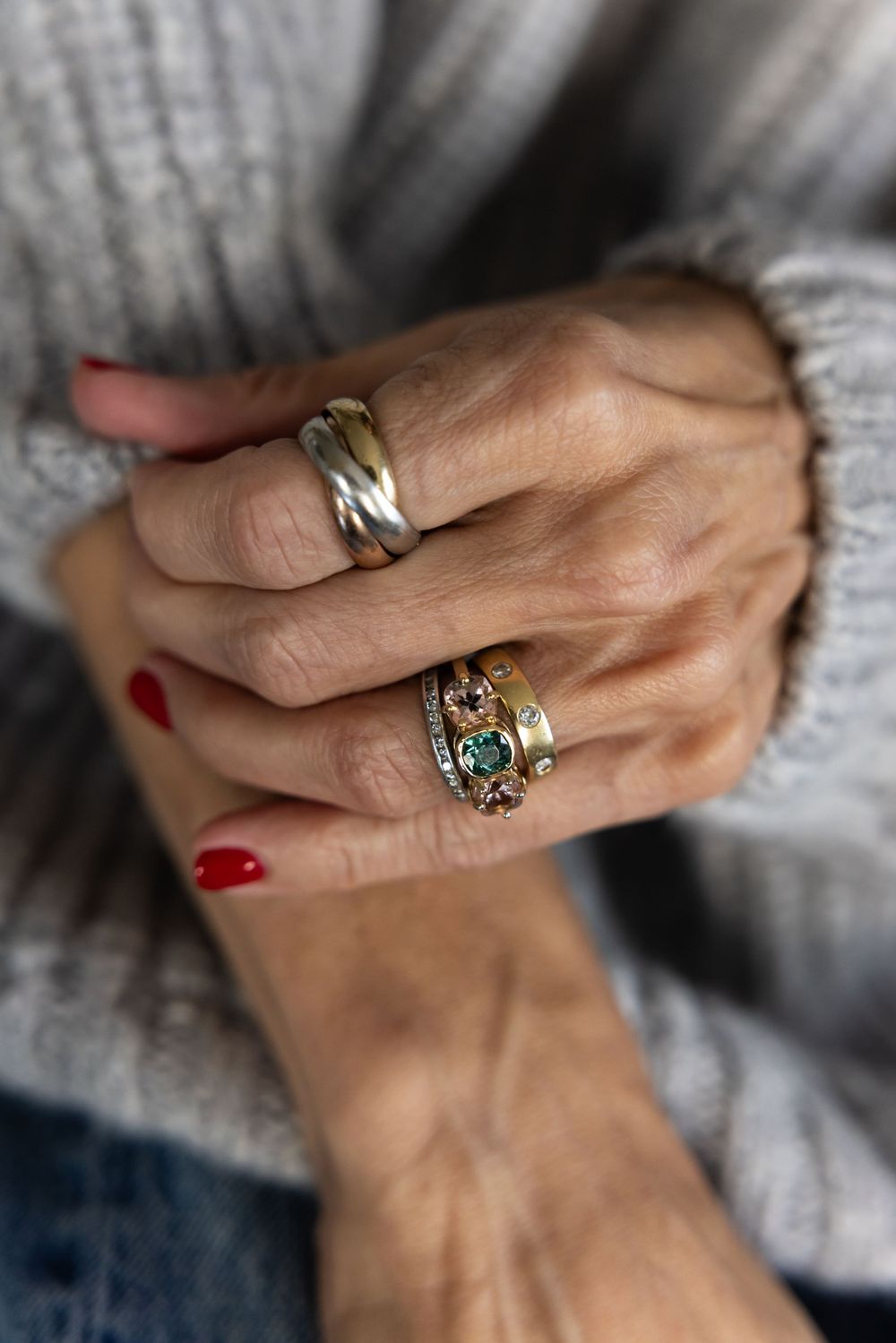 Close-up of folded hands adorned with gold and silver rings over a gray sweater.