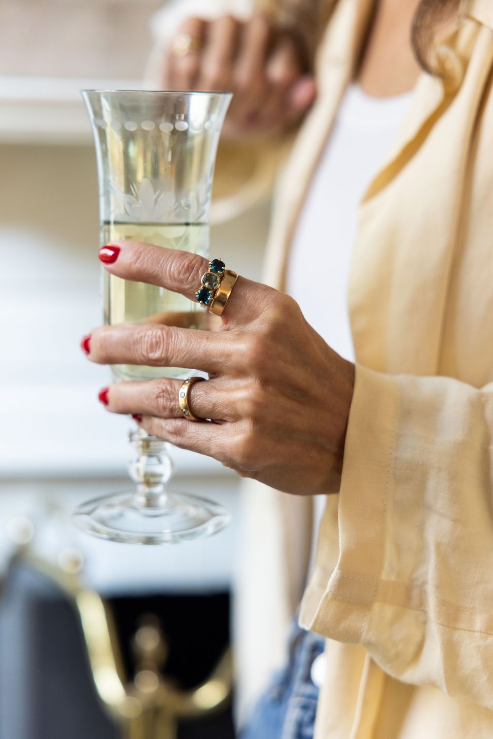 Woman holding a champagne flute with red nails and gold rings, wearing a beige blazer.