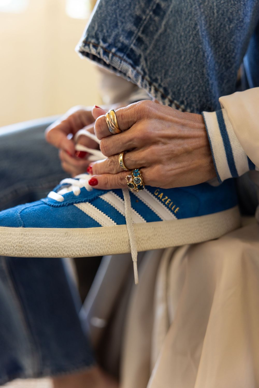 Close-up of hands with red nails and gold rings tying laces on blue Adidas Gazelle sneakers.