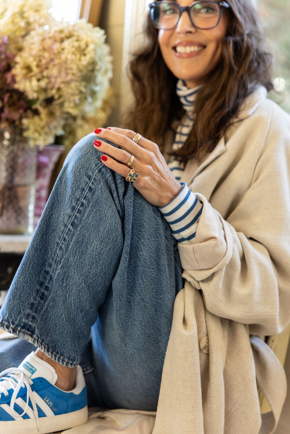 Smiling woman in glasses, beige coat, jeans, and blue Adidas Gazelle sneakers sitting relaxed.
