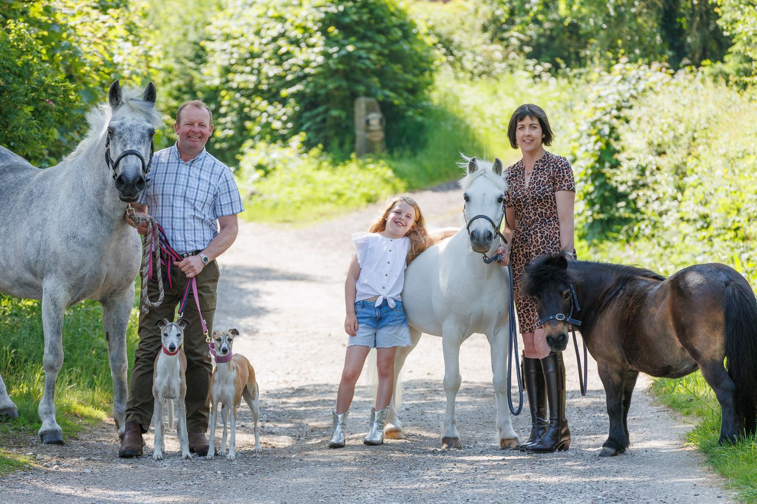 Family with two adults and a girl posing on a country path with three horses and a dog surrounded by greenery.