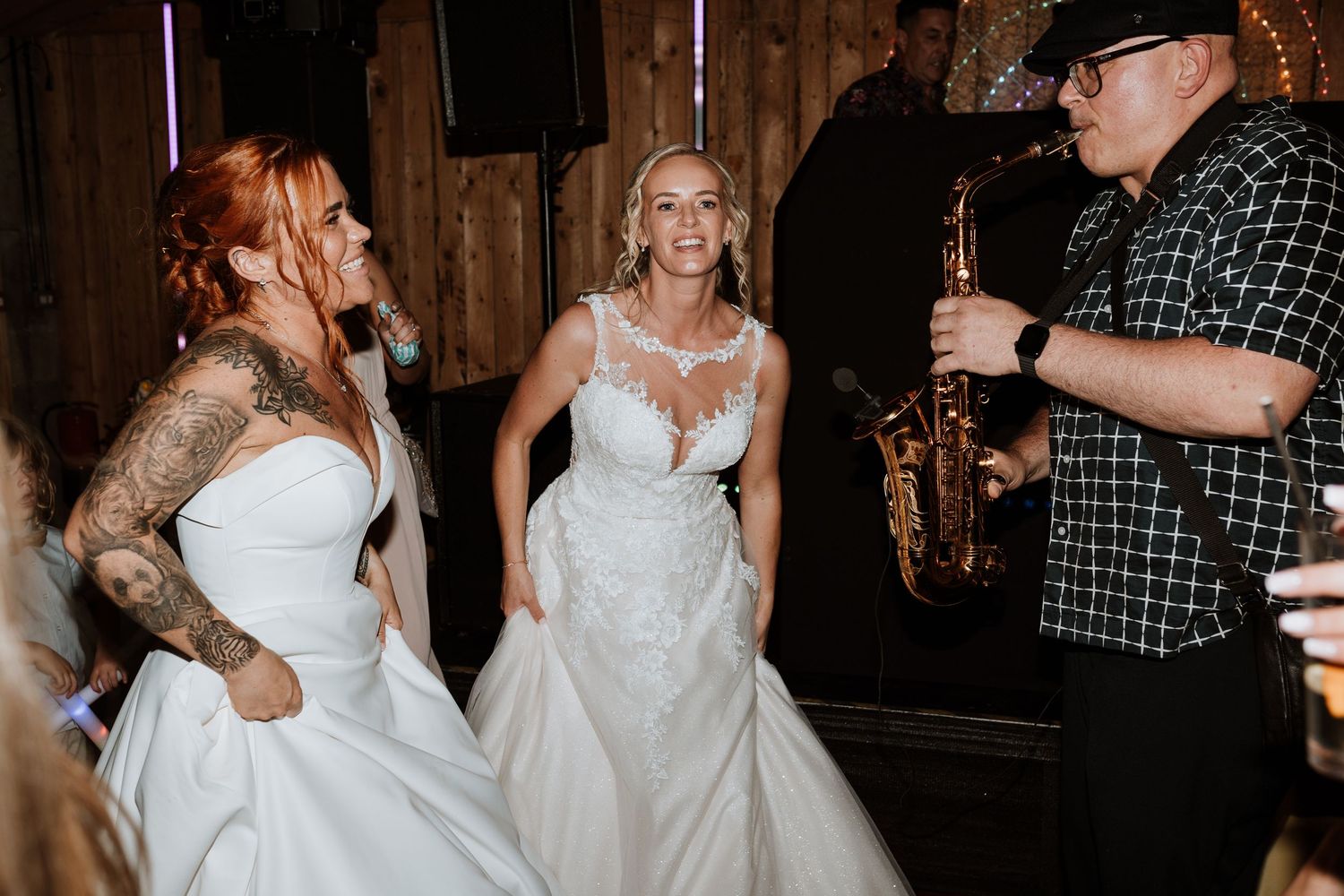 Two brides share first dance in rustic barn venue with warm lighting and rainbow arch backdrop.