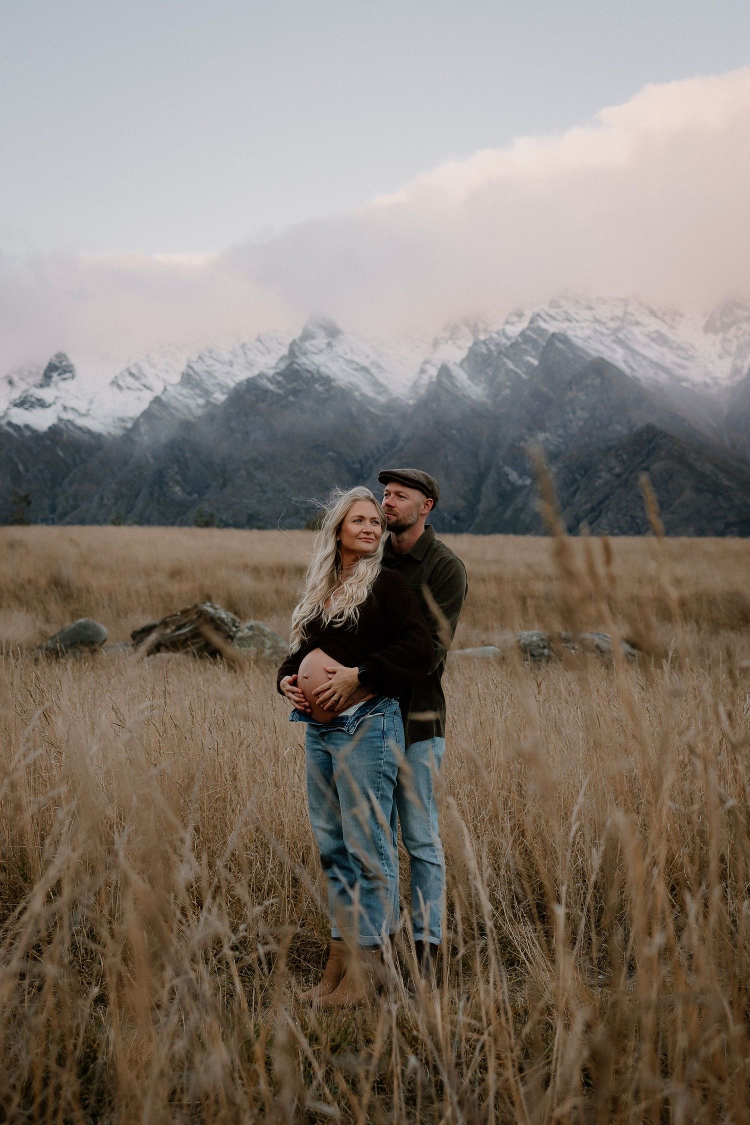 Couple embracing in golden field with snow-capped mountains backdrop at dusk.