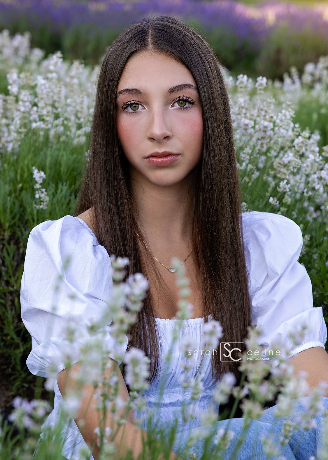 Young brunette woman in white dress poses gracefully among purple lavender flowers in a lush garden setting.