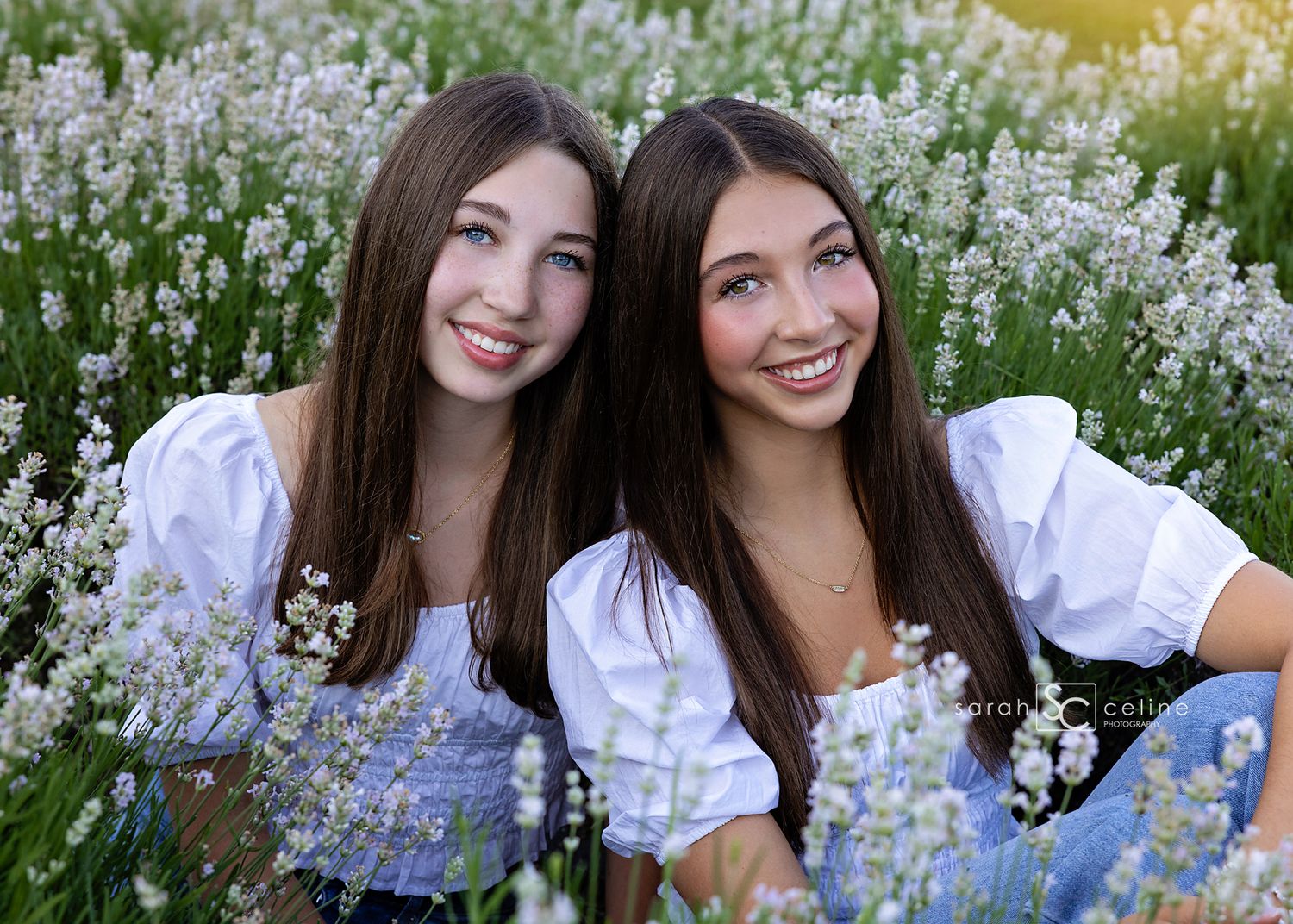 Two smiling teenage girls with long brown hair pose together in a blooming lavender field wearing white tops.
