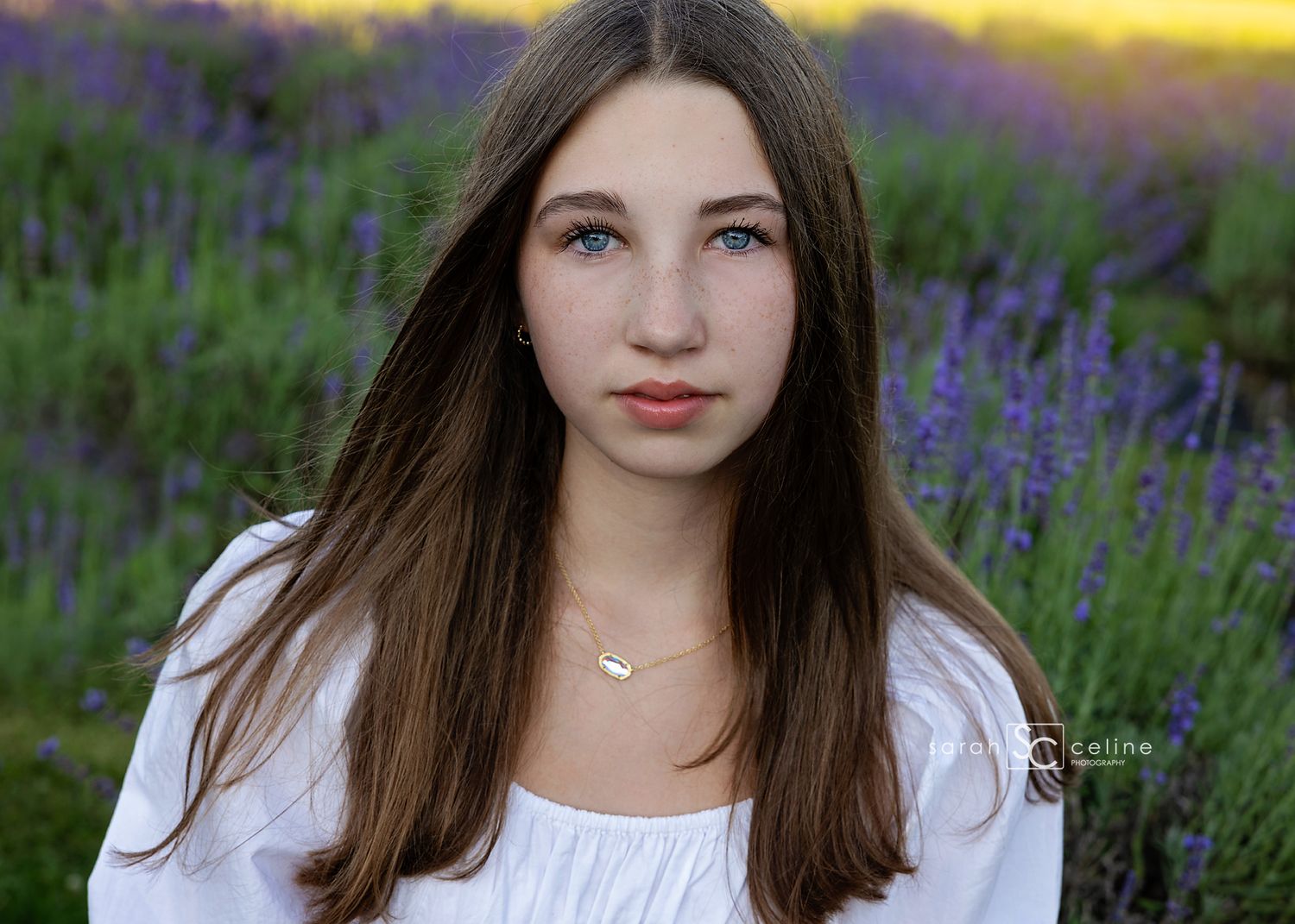 Teen girl with long brown hair poses in a lavender field wearing a white top, captured in golden hour light.