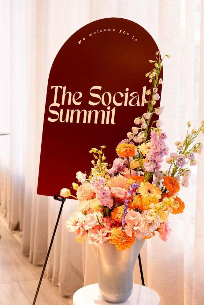 Elegant event welcome sign reading The Social Summit on a dark red arch display beside a vibrant floral arrangement in white vase.