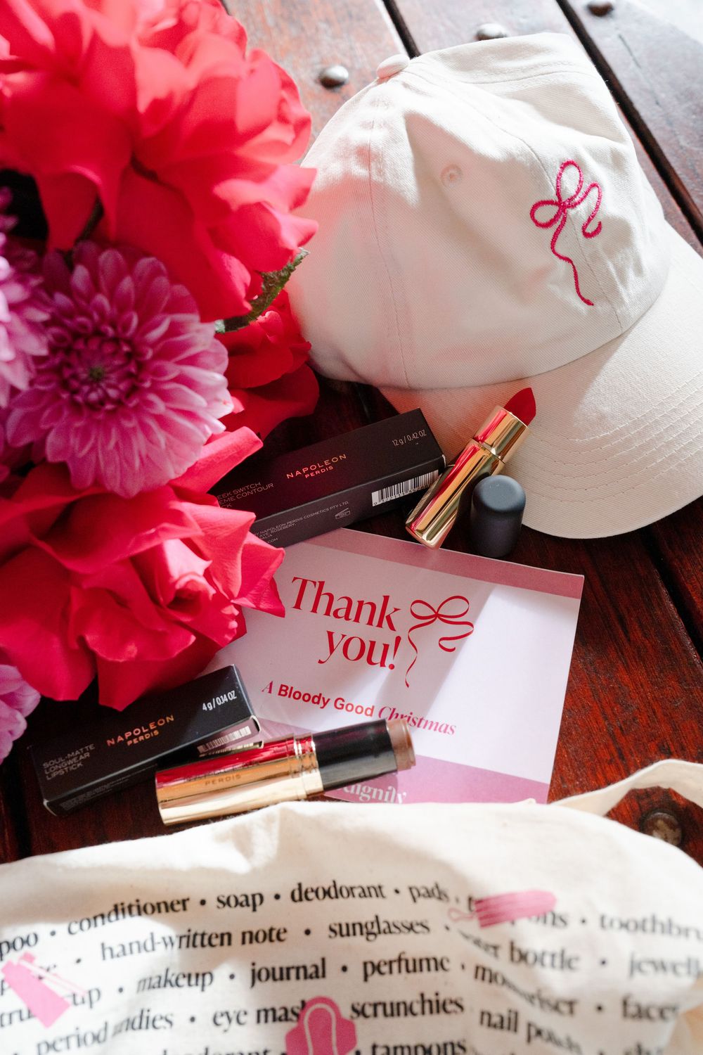 Pink flowers, makeup products, white cap with bow, thank you card, and tote bag arranged on wooden surface.
