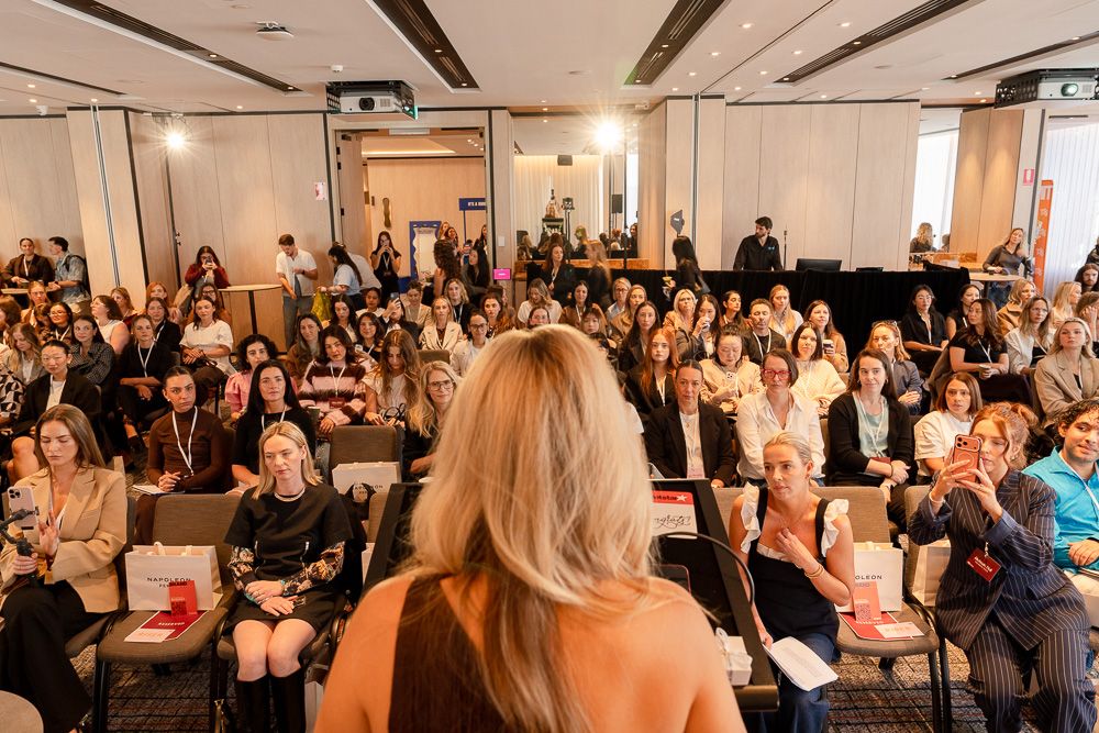 A female speaker addresses a large engaged audience at a professional women's conference in a modern venue.