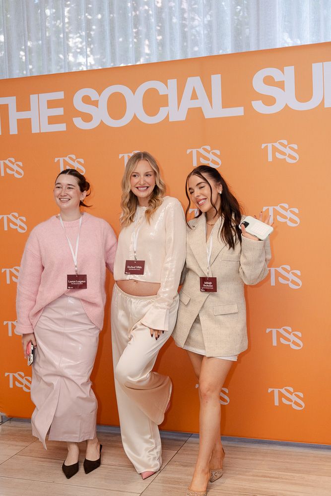Three women smiling at The Social Summit event, wearing neutral tones, posing in front of an orange TSS branded backdrop.