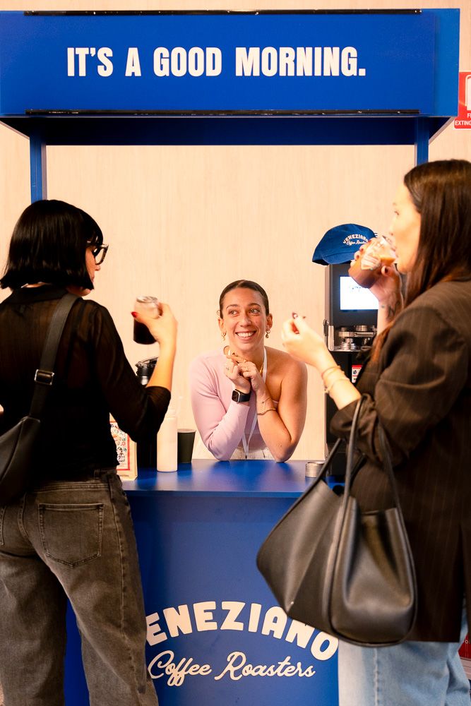 Smiling barista at Veneziano Coffee Roasters blue stand serving two women customers at a morning coffee event.