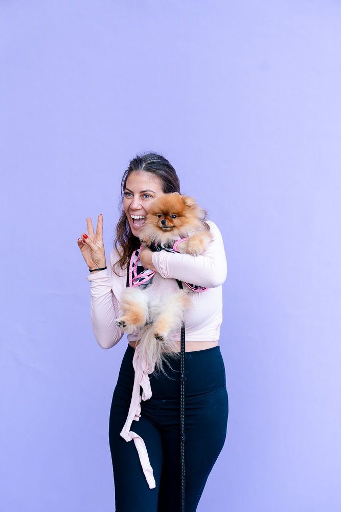 Woman smiling and making peace sign while holding a fluffy Pomeranian dog against a purple background.