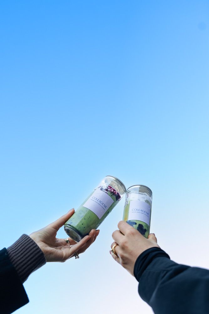 Two hands holding green matcha drinks in clear cups, toasting against a bright blue sky backdrop.