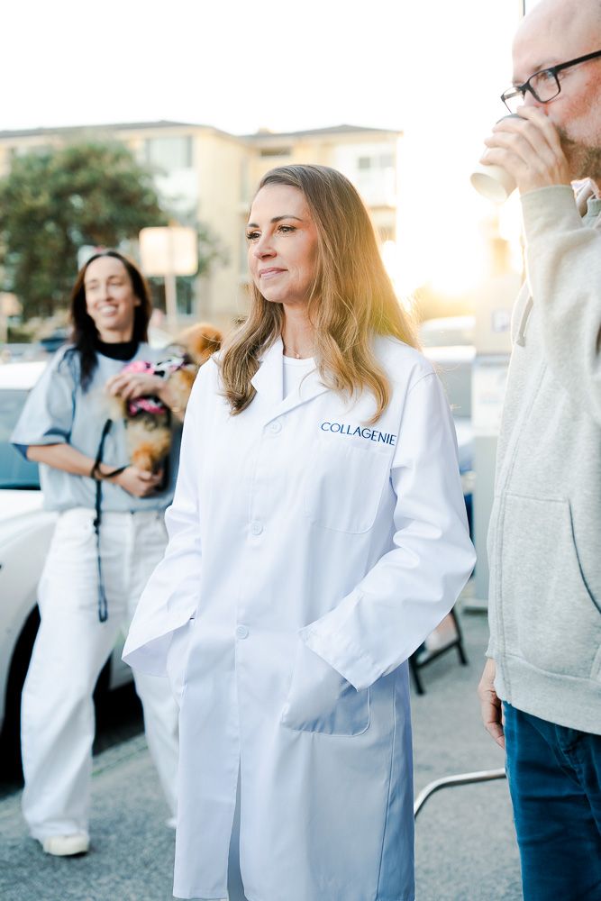 Woman in white Collagenie lab coat standing outdoors at golden hour with people and a dog in background.
