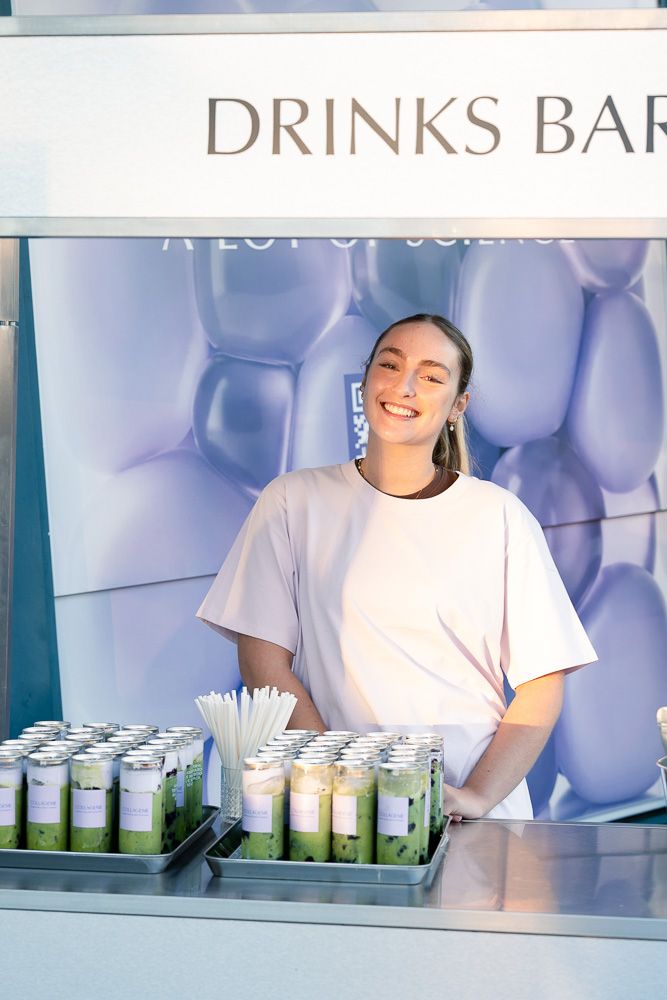 Smiling woman in white t-shirt standing at a drinks bar with green matcha beverages on display.