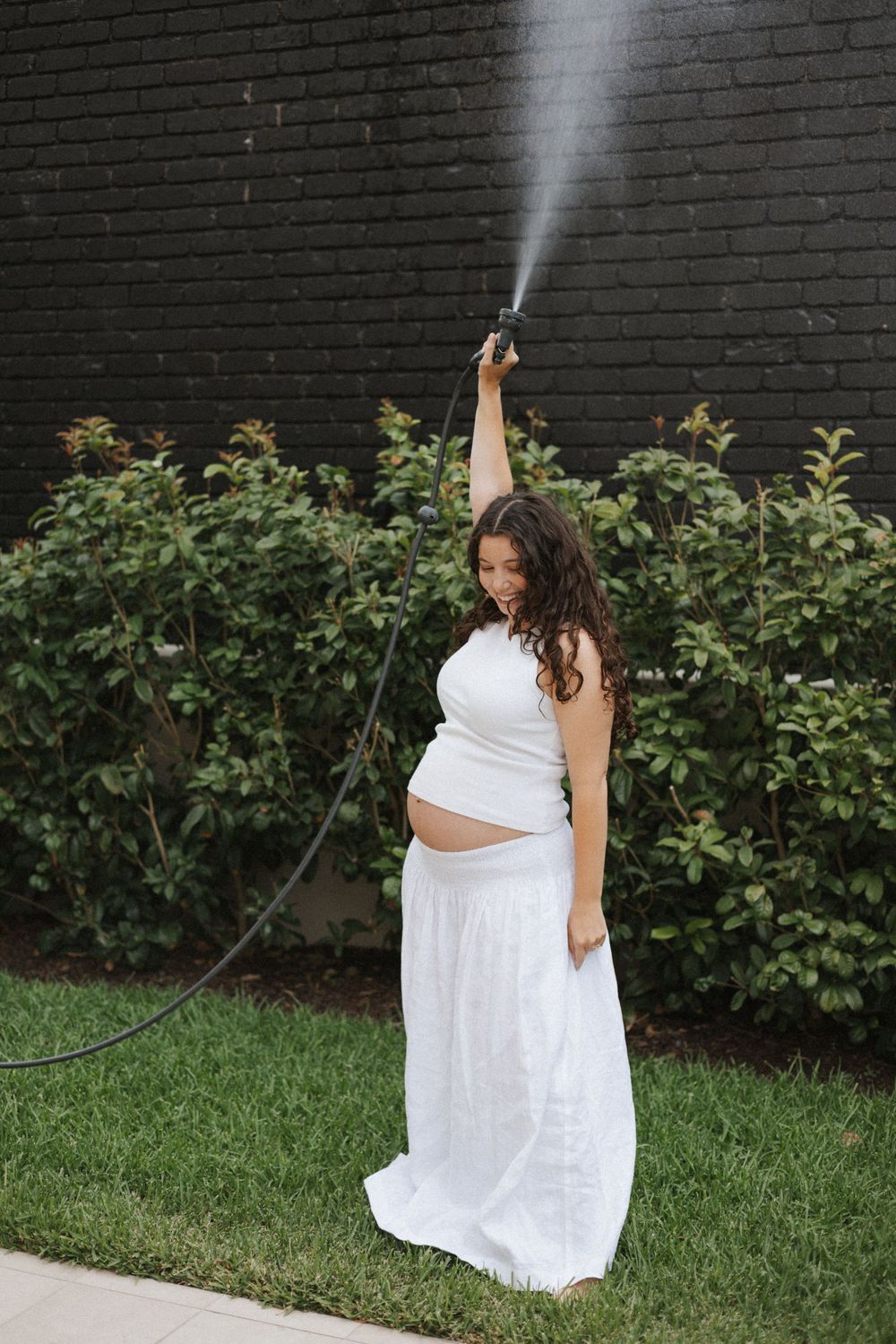 Pregnant woman in white outfit playfully spraying water hose overhead in a lush green backyard.