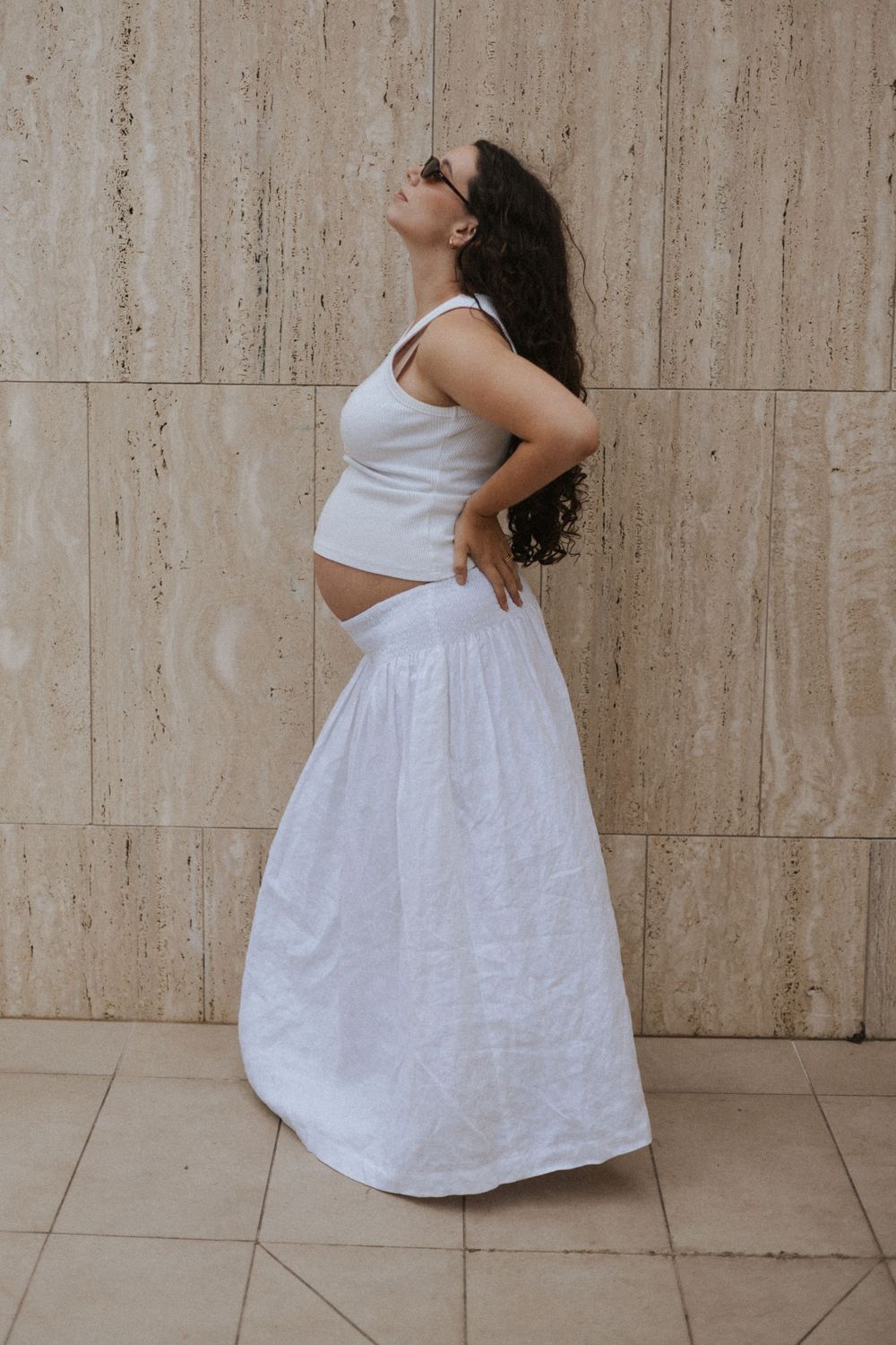Pregnant woman in white crop top and maxi skirt posing sideways against a travertine wall.