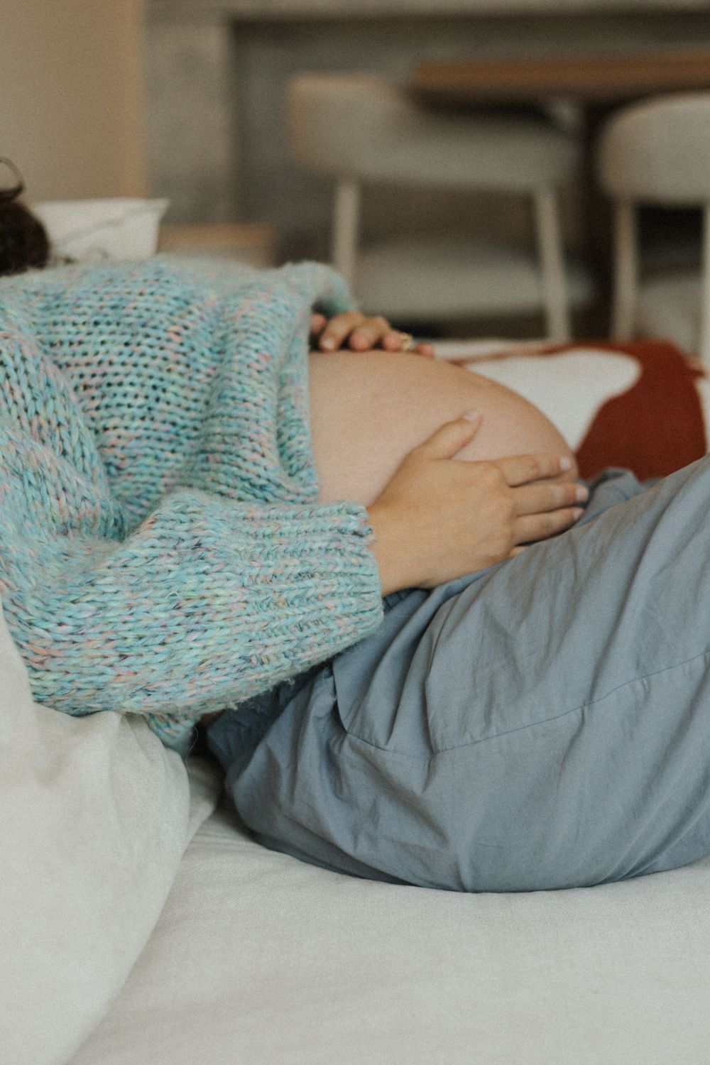 Pregnant woman in teal knit sweater lying on bed gently holding her bare baby bump.
