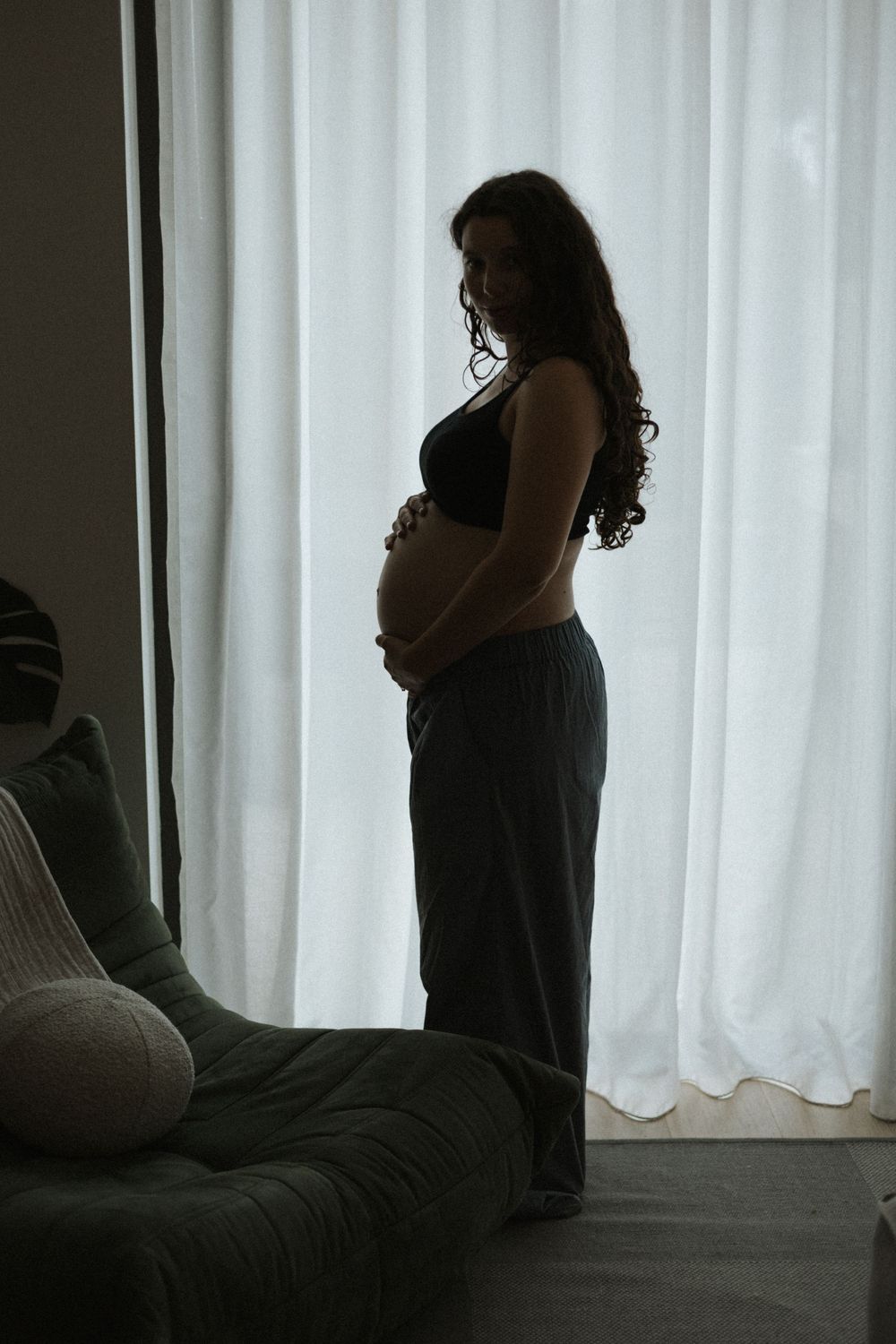 Silhouette of a pregnant woman holding her belly standing in front of sheer white curtains.