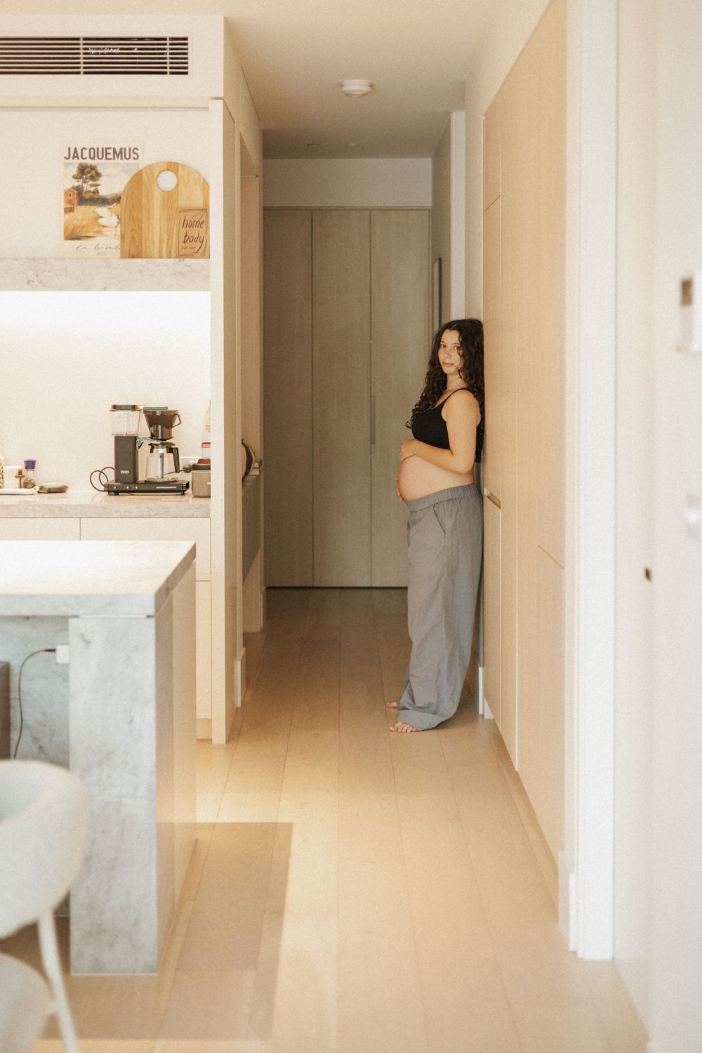 Pregnant woman in black crop top and gray pants standing in a modern apartment hallway.