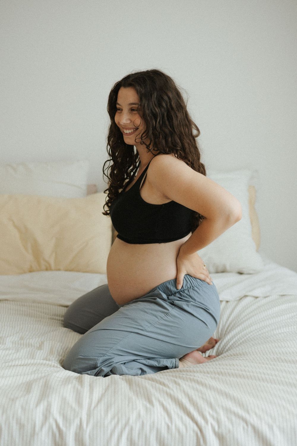 Smiling pregnant woman in black crop top and gray pants kneeling on a white bed.