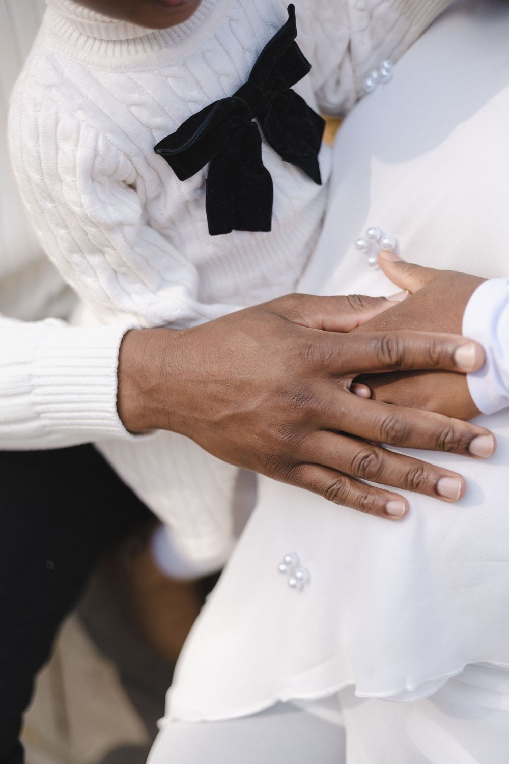 Close-up of a couple holding hands in white outfits, with a black bow tie accent, symbolizing love and togetherness.