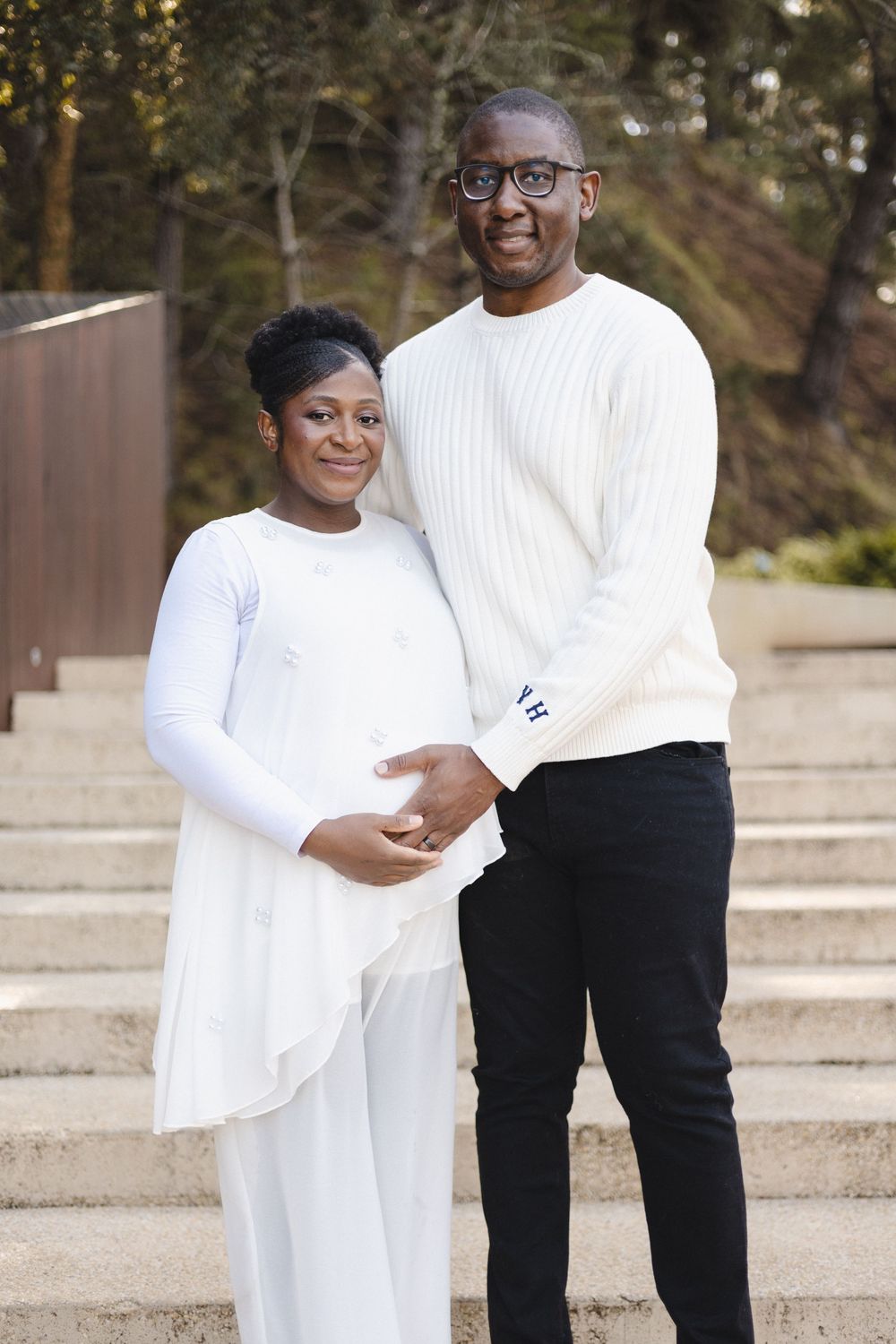 A couple in matching white outfits poses on outdoor steps, the woman visibly pregnant as the man tenderly holds her belly.
