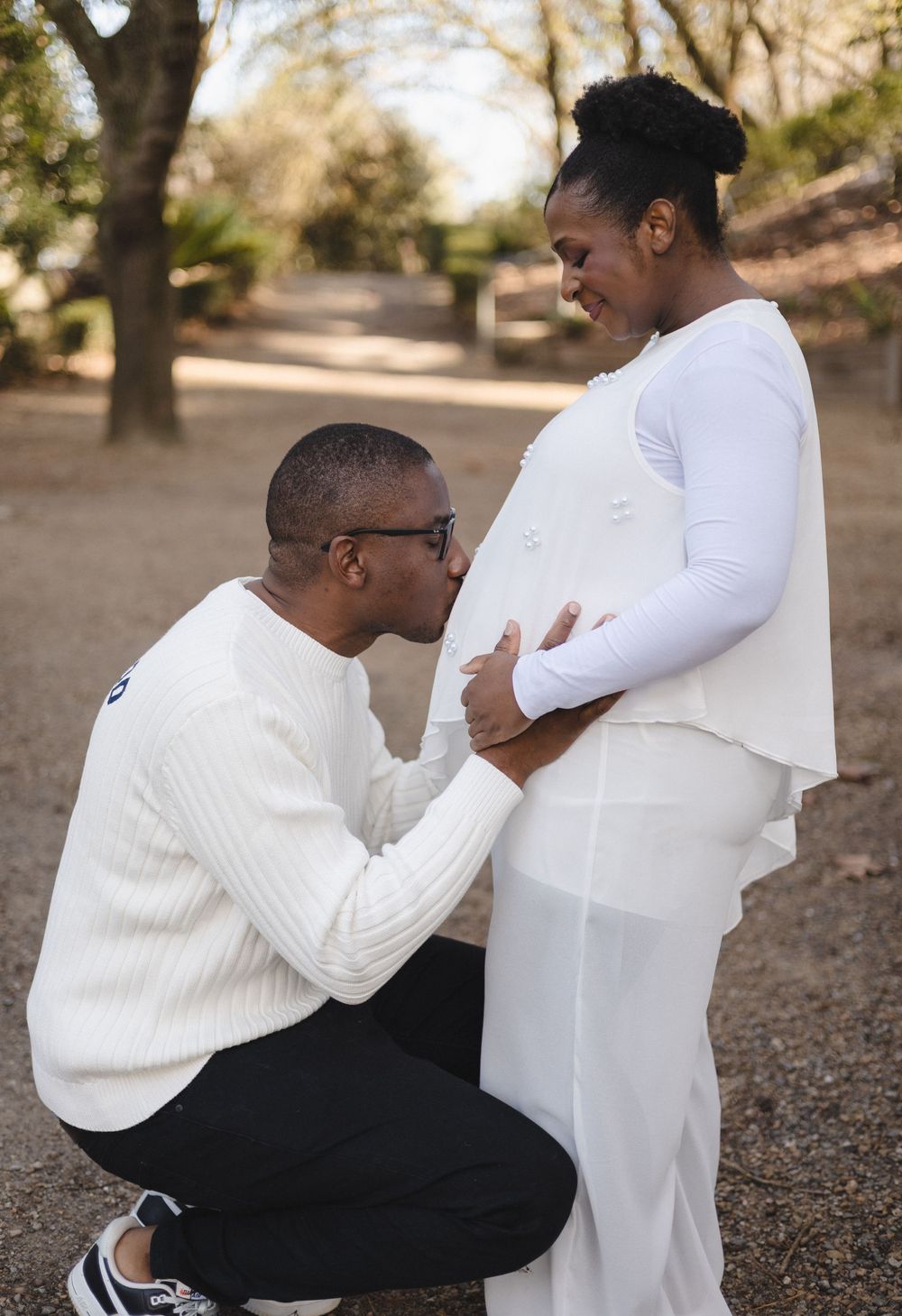 A man kneels kissing a pregnant woman's belly outdoors, both dressed in white, in a tender maternity photo shoot.