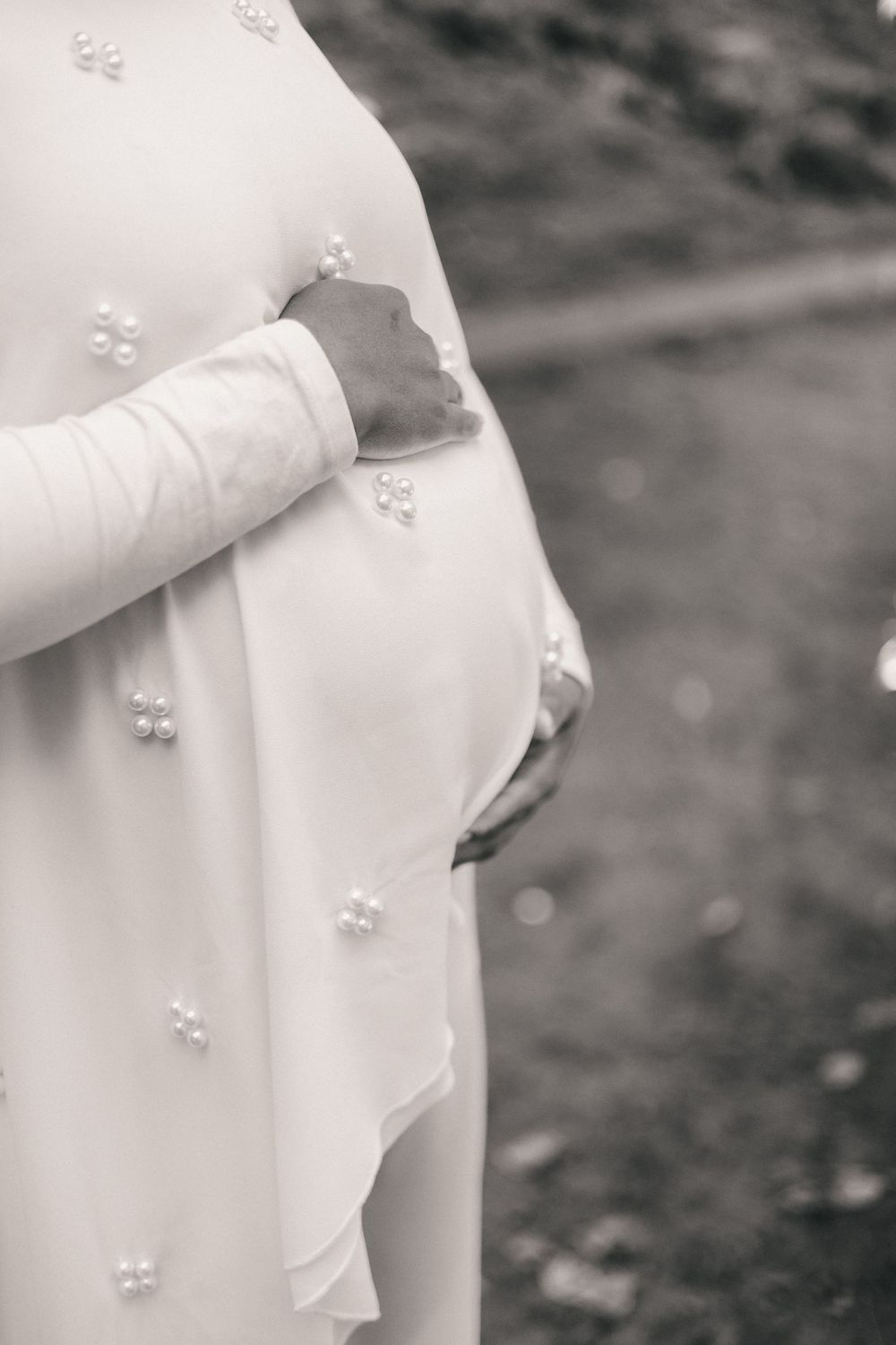Black and white photo of a pregnant woman in a pearl-embellished white dress cradling her baby bump outdoors.