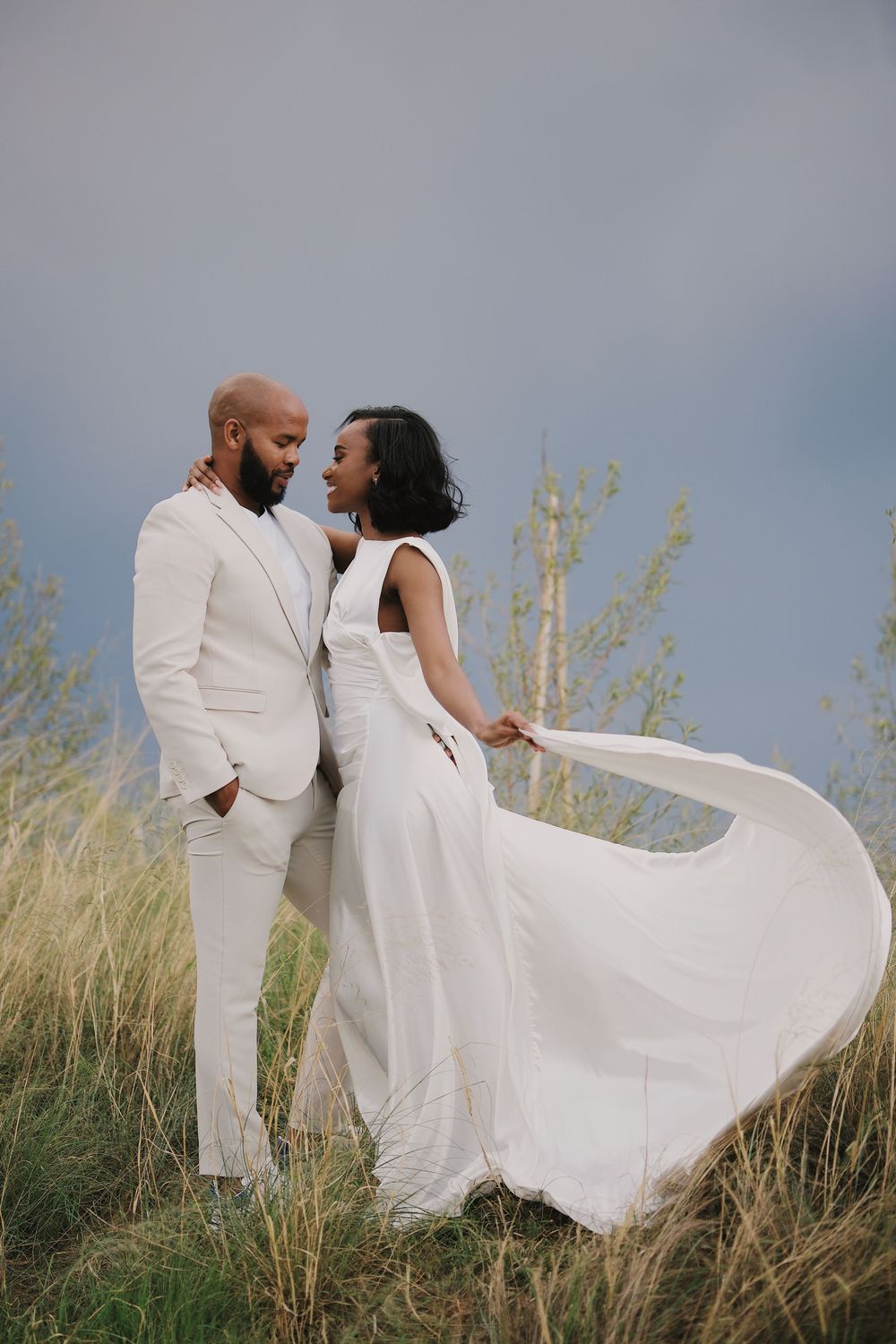 Couple in wedding attire embrace in golden field, bride in white flowing dress, groom in ivory suit under dramatic sky.
