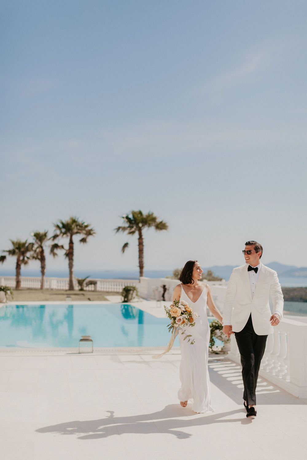 Wedding couple walks hand in hand beside infinity pool with palm trees and ocean view in sunny Mediterranean setting.