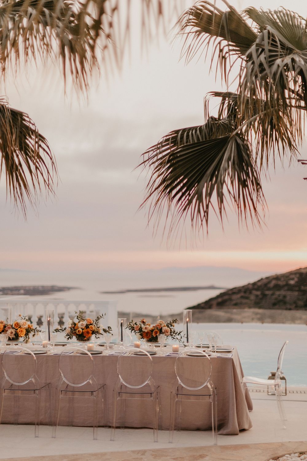 Romantic outdoor wedding reception table set under palm trees with ocean views and a pastel sunset sky.