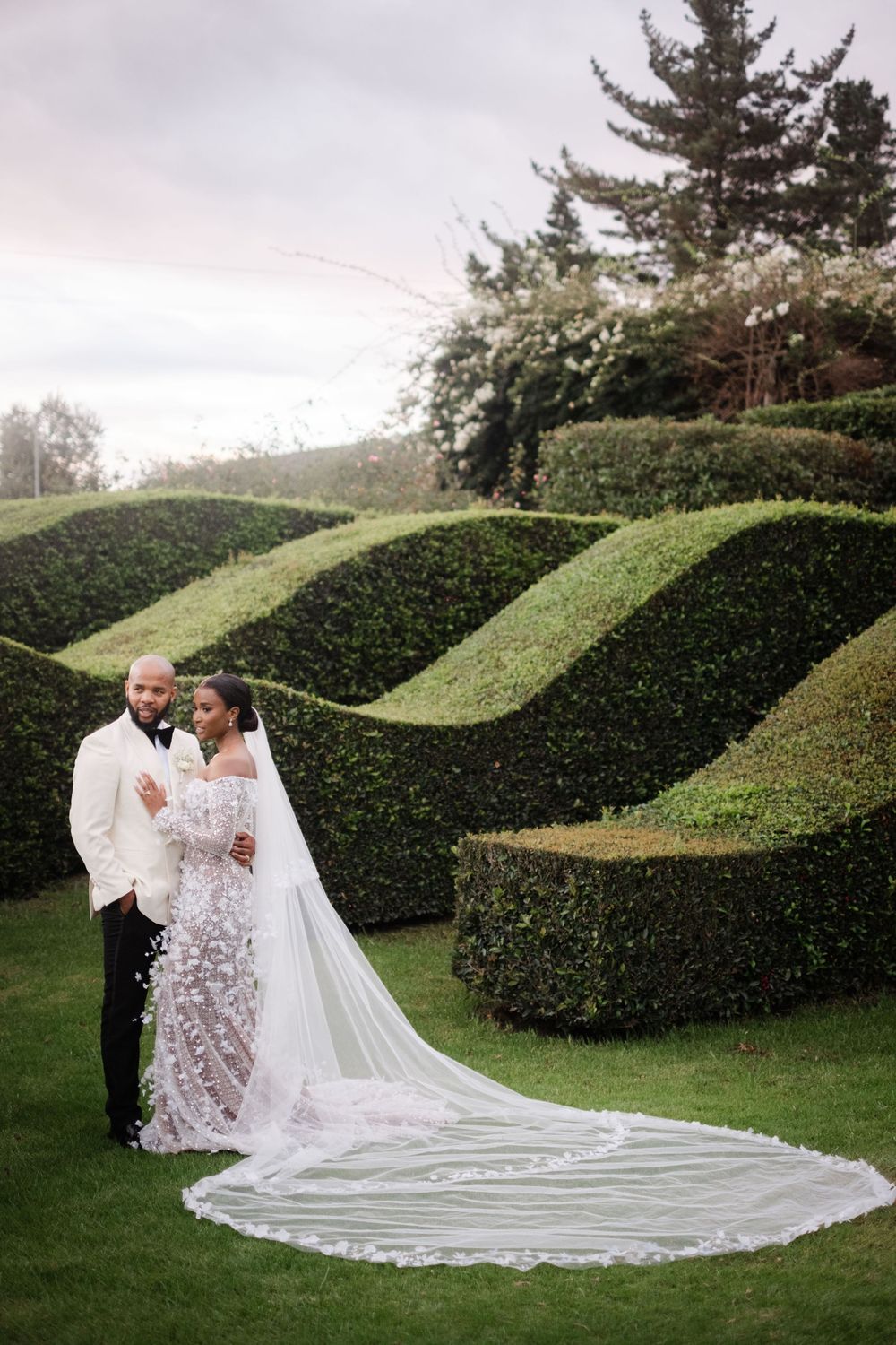 Bride and groom pose in lush garden with sculpted hedges, bride wearing lace gown with cathedral veil.