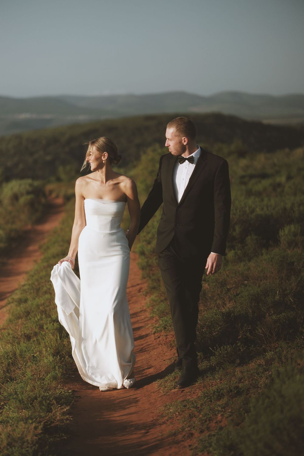 Bride and groom walking hand in hand along a scenic red dirt path at golden hour.