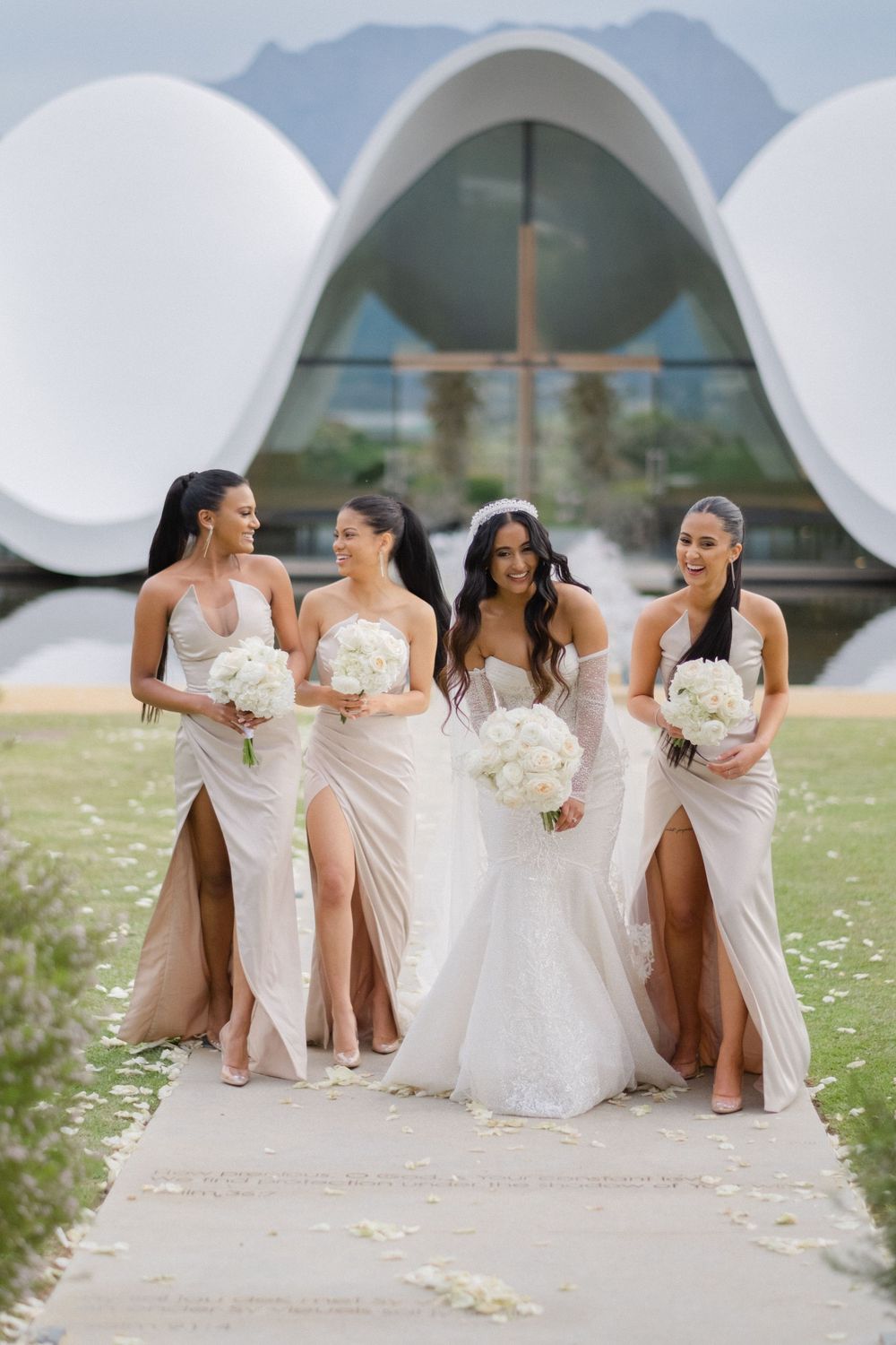 Bride walks with three bridesmaids in champagne gowns holding white bouquets at modern venue.