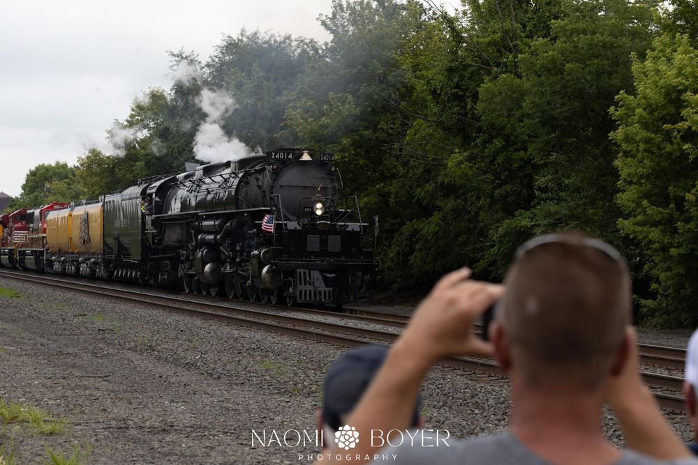 Man photographs a massive black steam locomotive billowing smoke on railroad tracks surrounded by green trees.