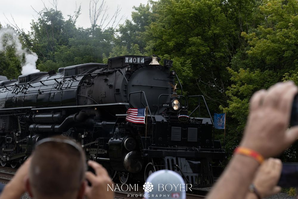 Crowds photograph a massive black steam locomotive with American flags, billowing smoke, at an outdoor railway event.