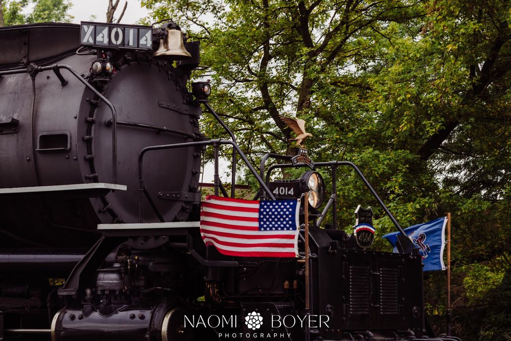 Vintage steam locomotive X4013 adorned with American flag, surrounded by lush green trees, with crew members visible on board.