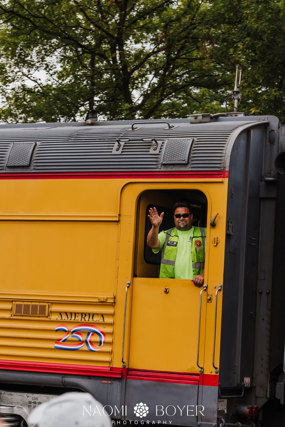 A smiling railroad worker in a safety vest waves from the window of a yellow Union Pacific America 250 train.