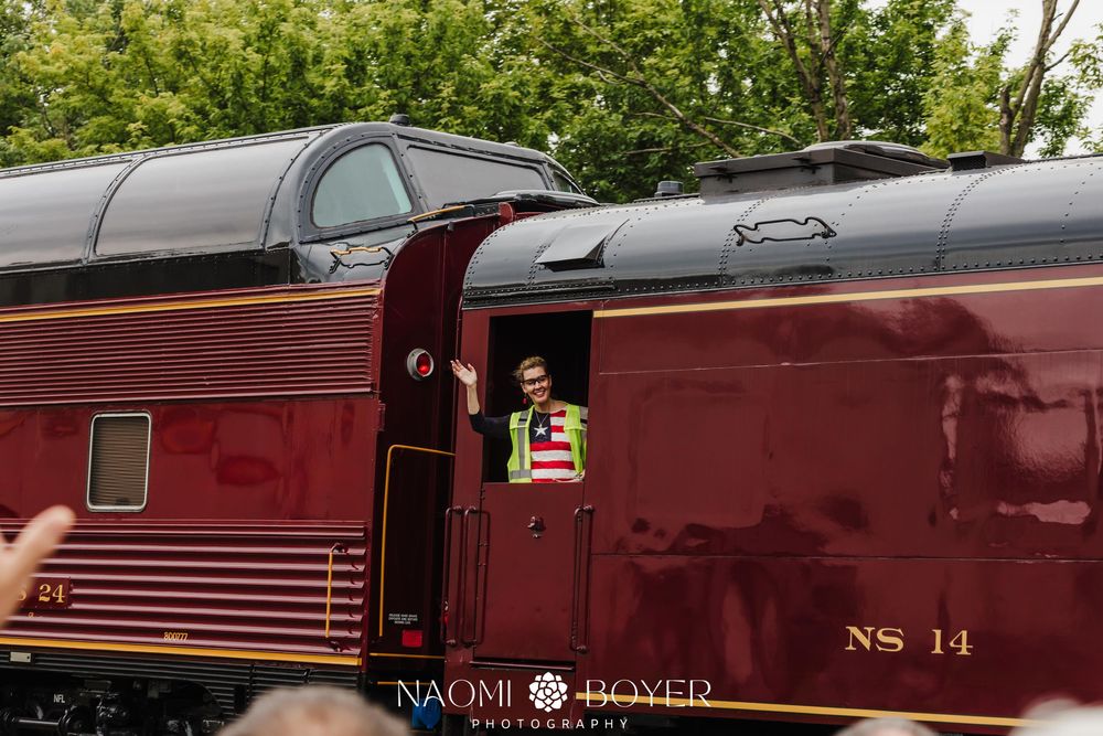A person waves from the doorway of a maroon NS 14 passenger rail car during a train event.