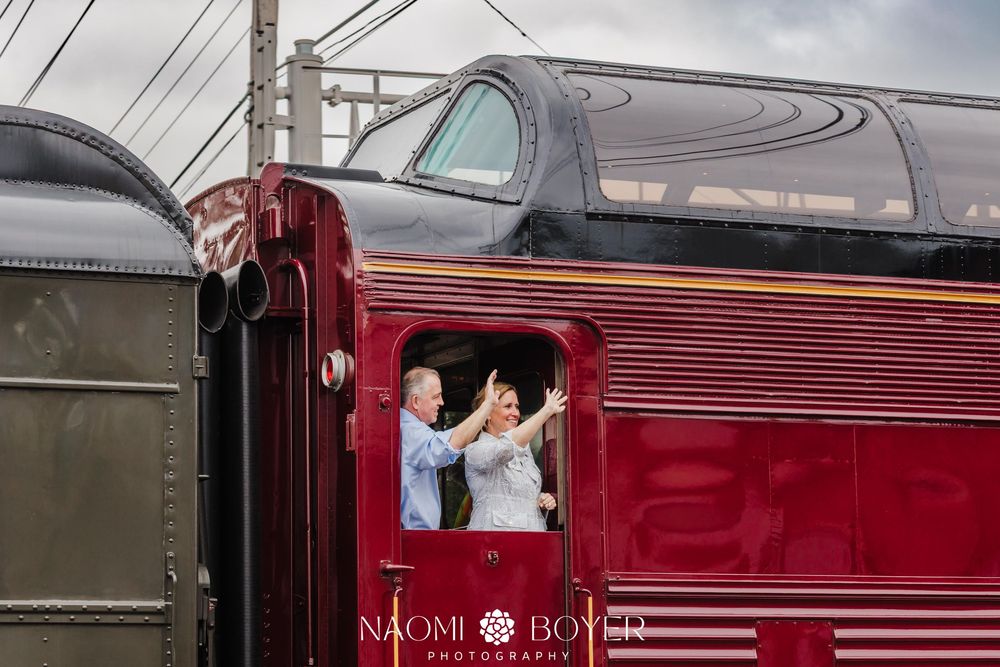 Two people wave from the window of a classic burgundy passenger train car on an overcast day.