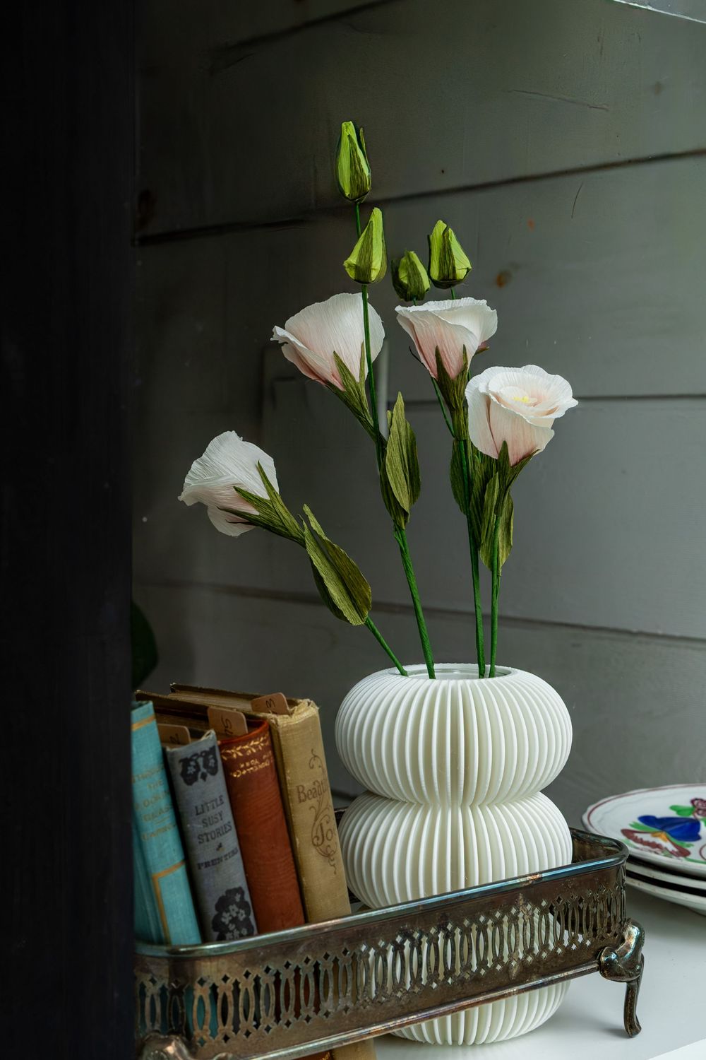 White ribbed vase with blush lisianthus flowers beside vintage books in an ornate metal tray on a shelf.Swell House La Grange